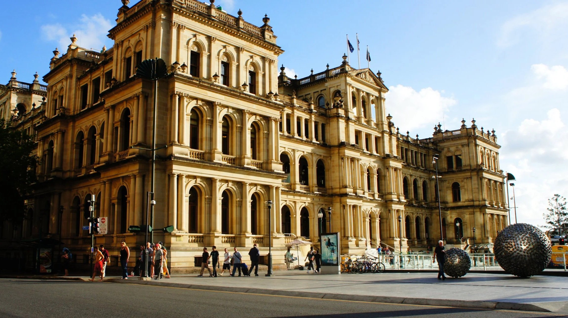 A historic, yellow sandstone building with classical architectural features, including arched windows, columns, and decorative balconies, under a bright blue sky with a few clouds, with people walking on the street in front.