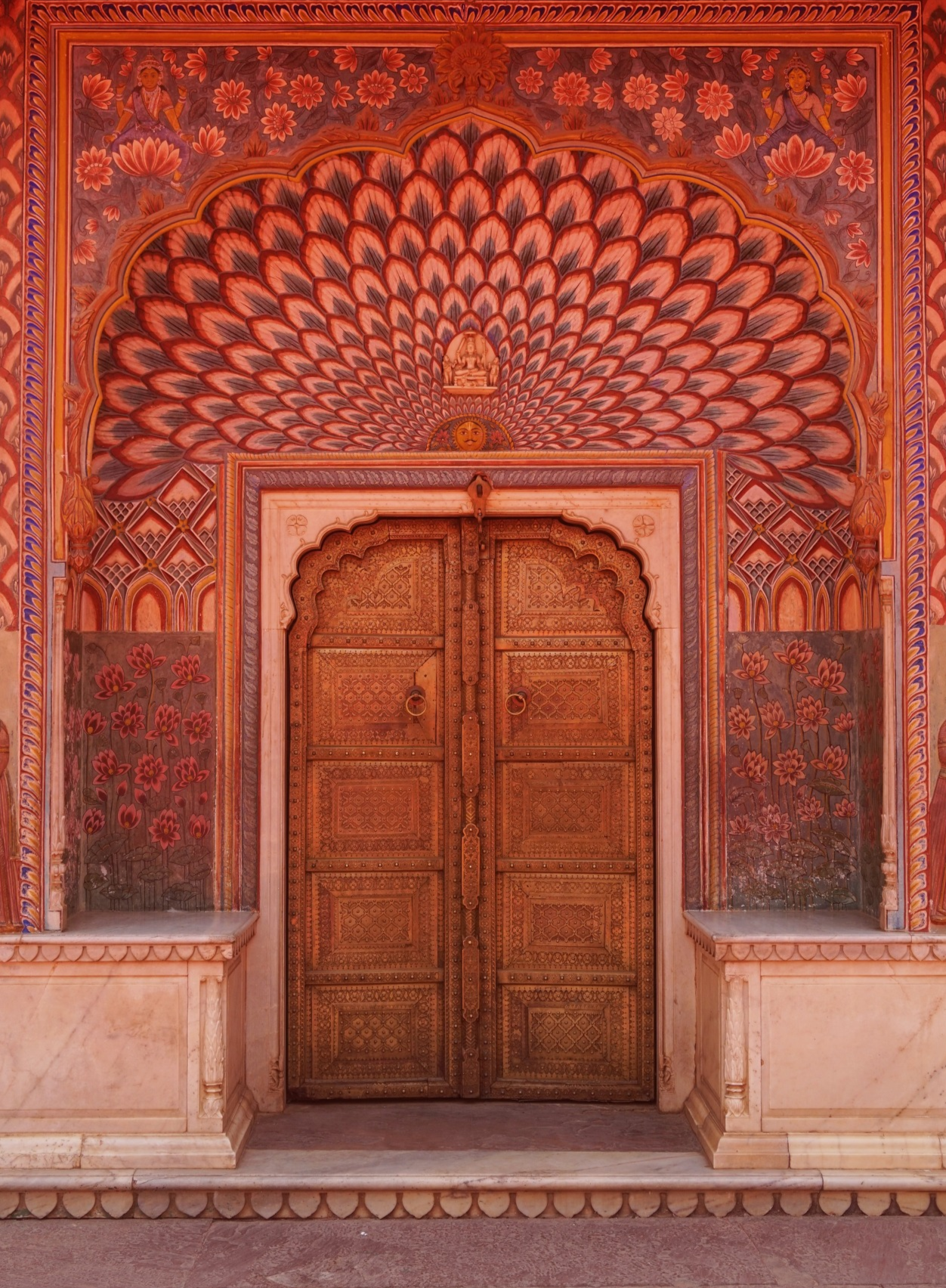 Intricate wooden front door with ornate carvings, flanked by floral and geometric murals, and a vibrant, peacock feather-like ceiling design.