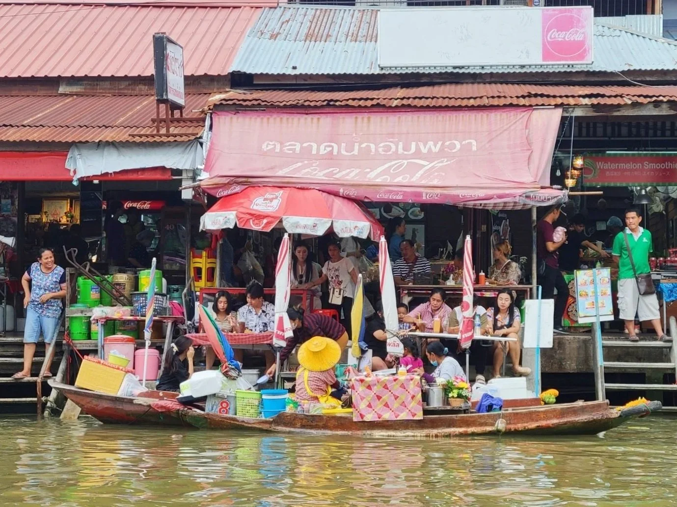People, including women and children, socializing and eating on a floating market boat near riverbank stalls with umbrellas and signs.