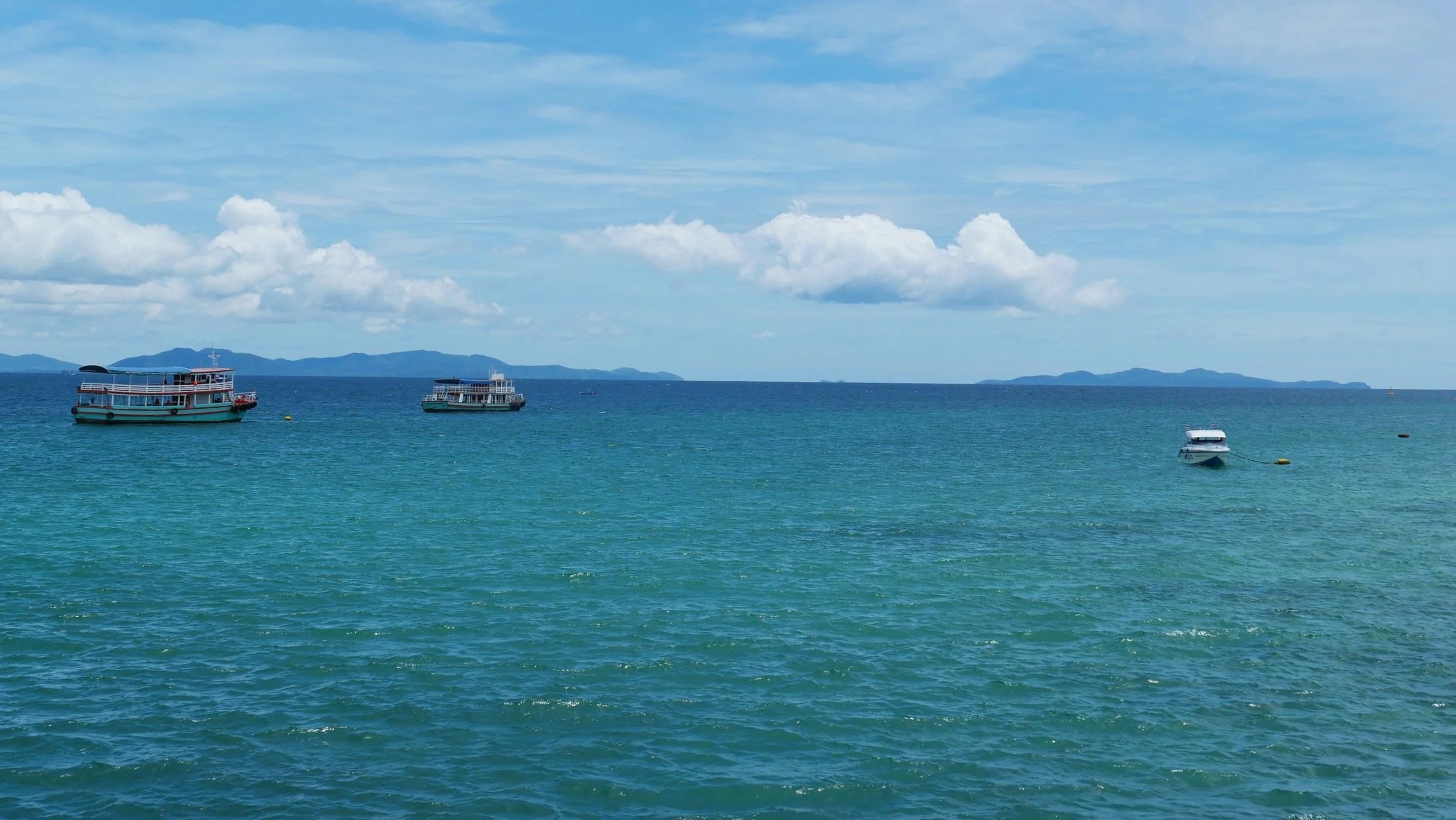 Boats floating on blue ocean water with distant islands and partly cloudy sky.