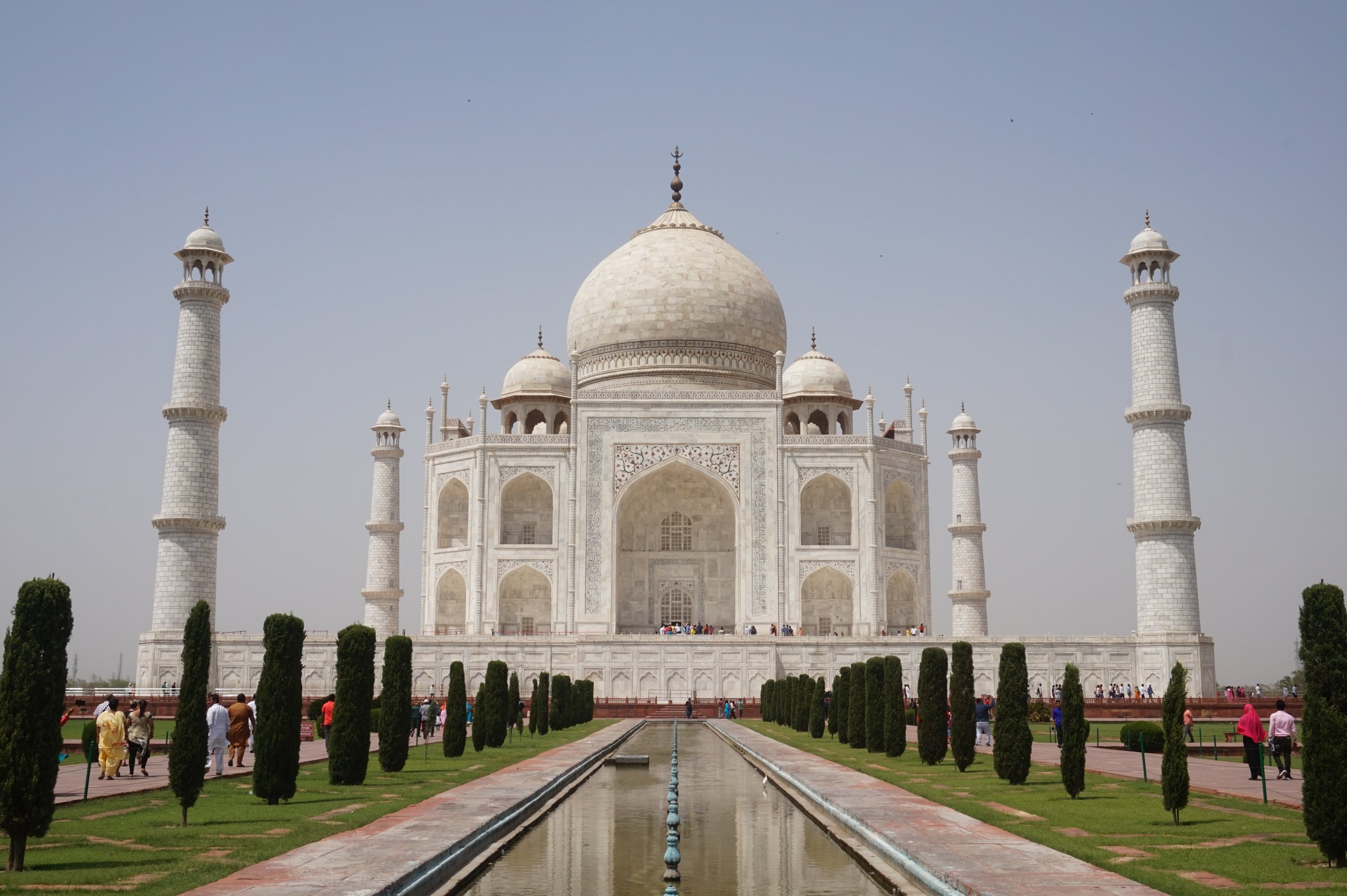 The Taj Mahal, a white marble mausoleum with a large central dome and four minarets, surrounded by a reflecting pool and lush gardens with stone pathways and green trees, under a clear blue sky.