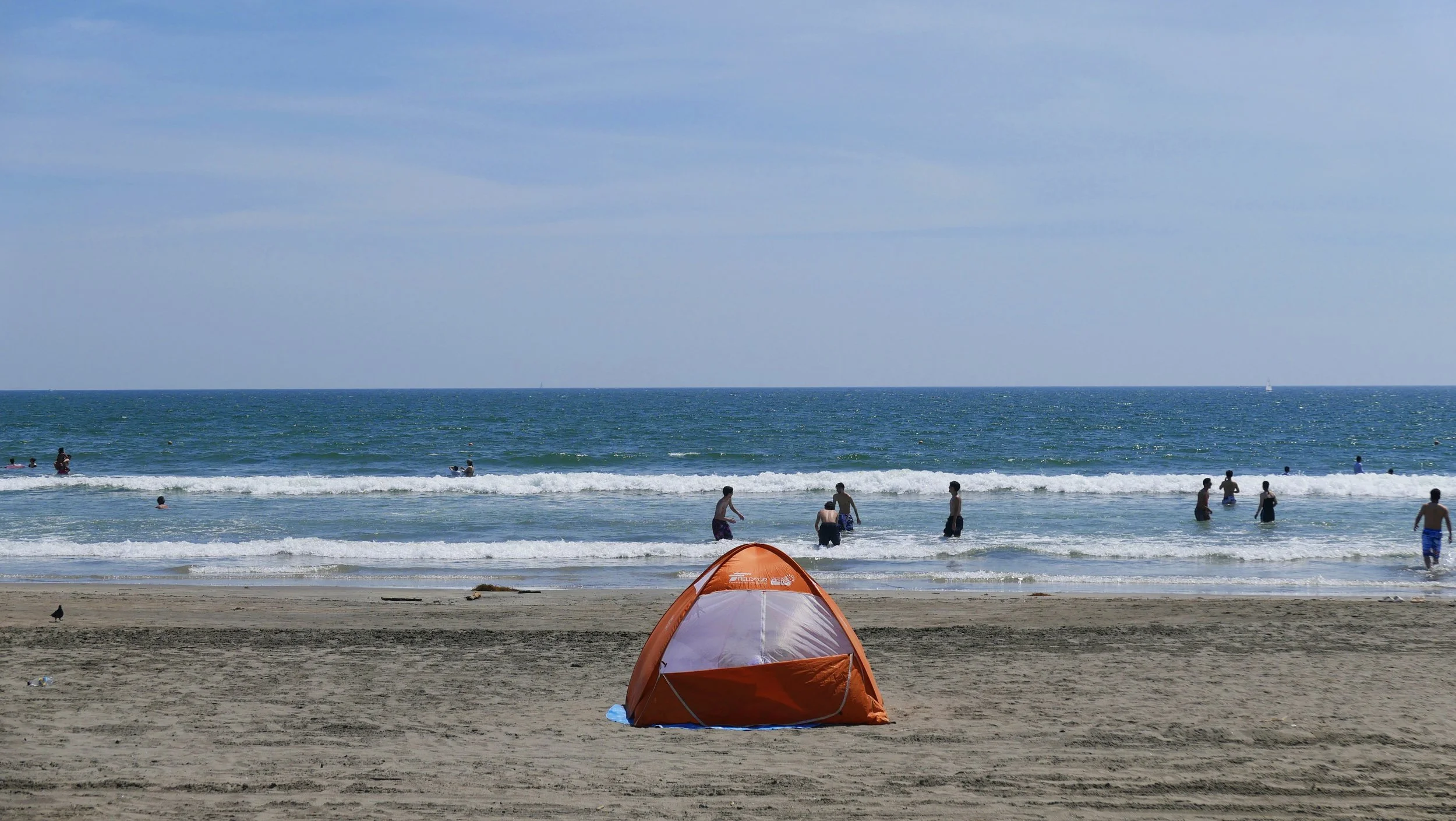 People swimming and playing in the ocean at a beach with cloudy sky, an orange tent on the sand in the foreground.