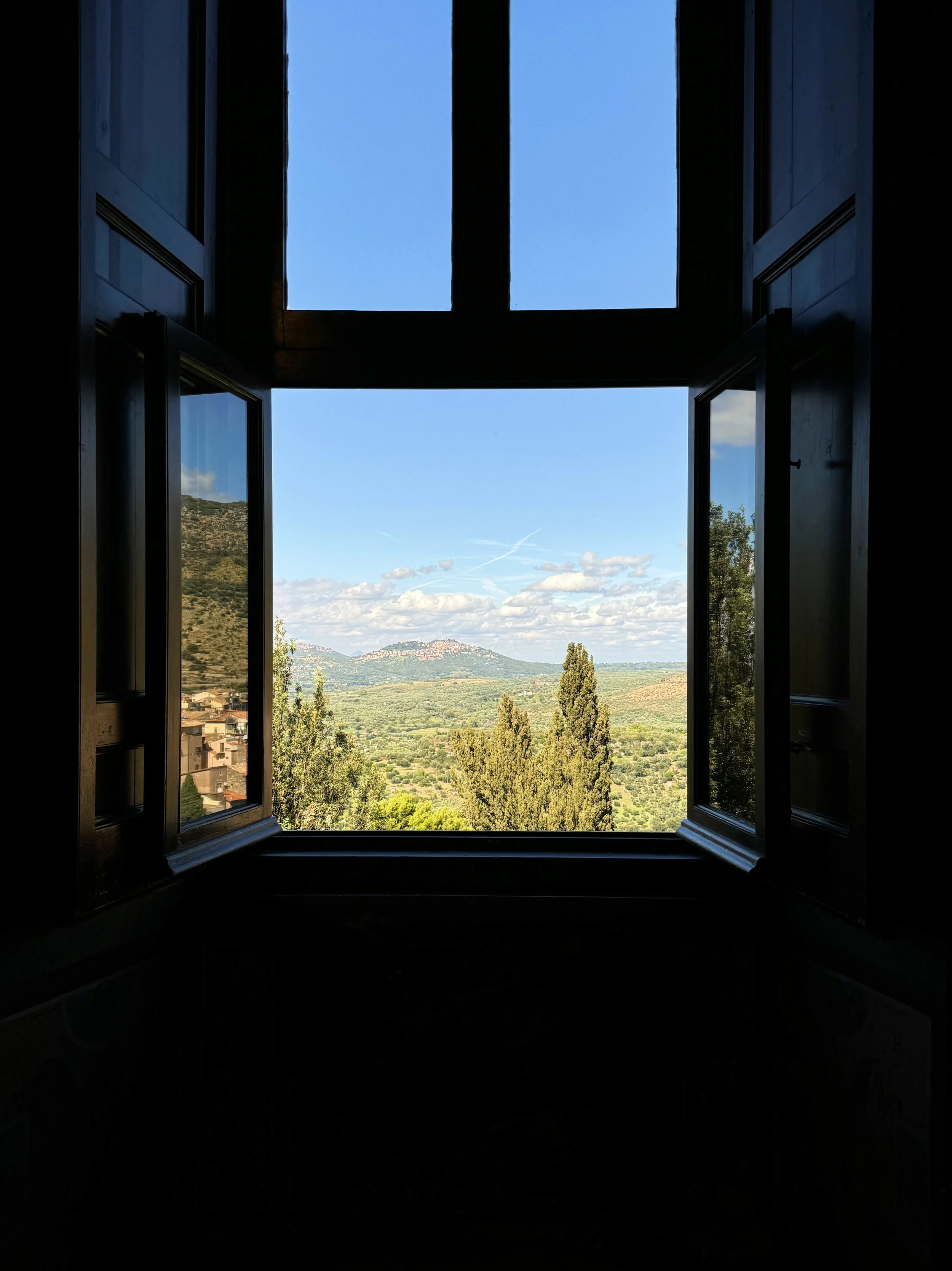 View through an open window showing distant mountains, trees, and a partly cloudy blue sky.