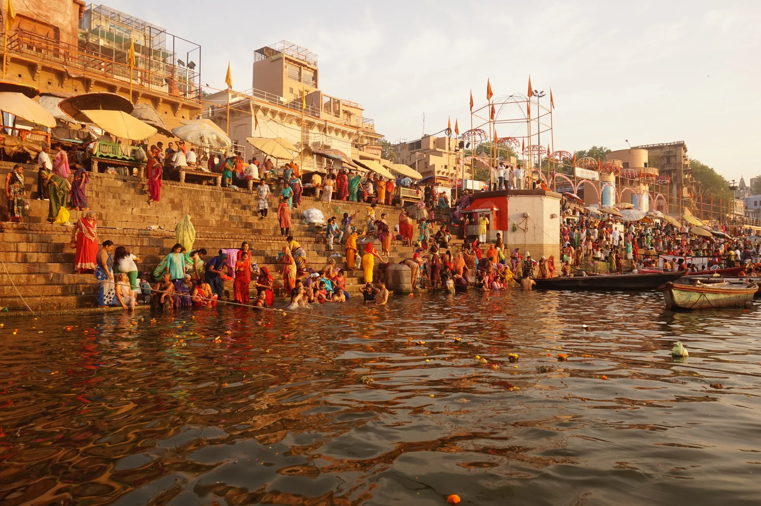 People gather on steps leading down to a riverbank, engaging in a ritual or festival, with boats in the water. The scene is set during sunset with warm lighting.