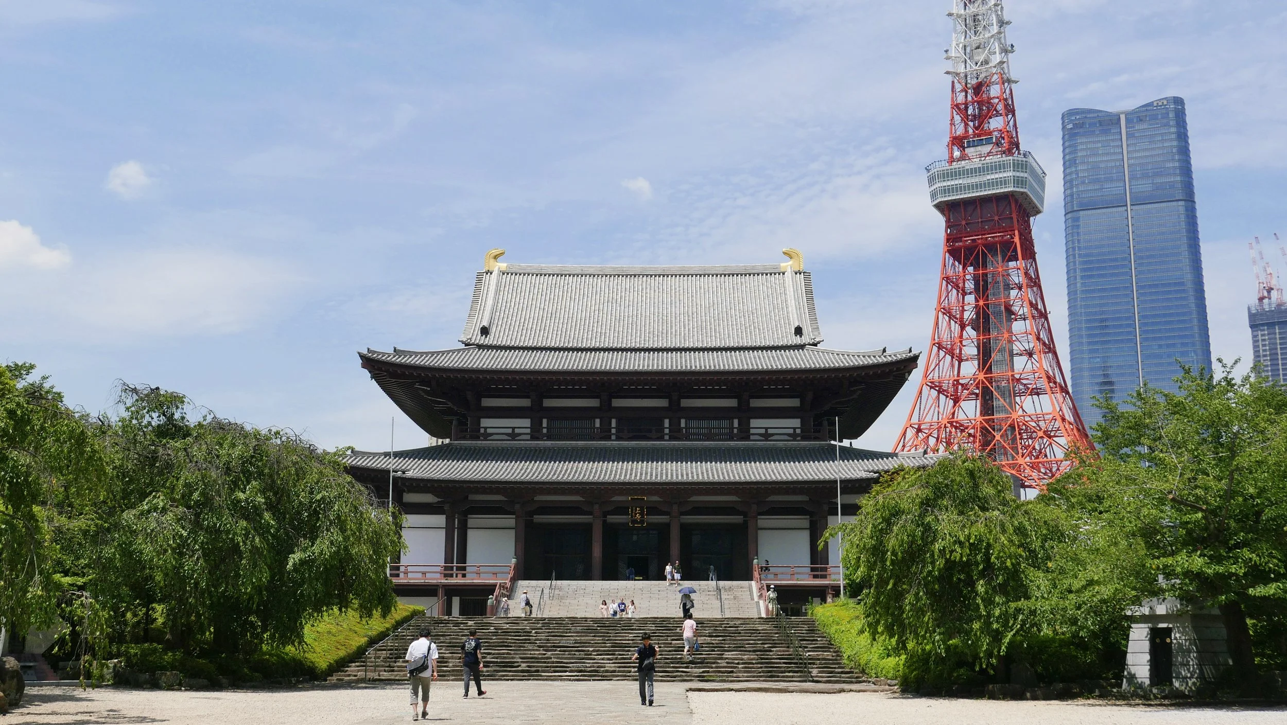 The image features a traditional Japanese temple with a pagoda-style roof, steps leading up to it, and surrounded by greenery, with the Tokyo Tower and modern skyscrapers in the background under a partly cloudy sky.