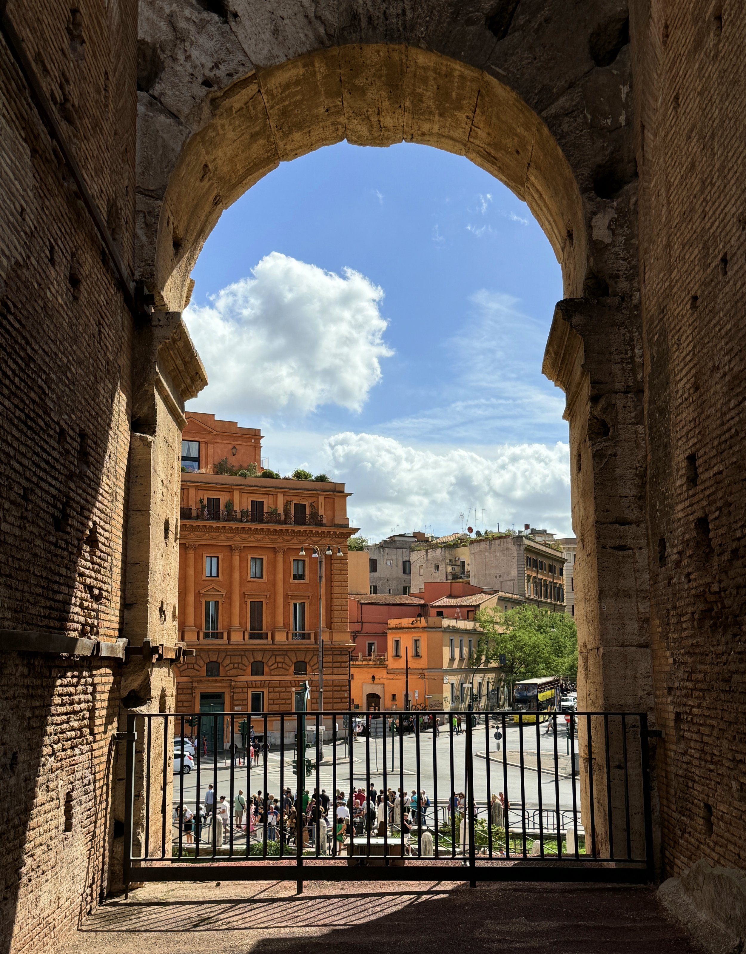 View of a city street and buildings through an ancient stone arch with a metal gate, under a blue sky with clouds.