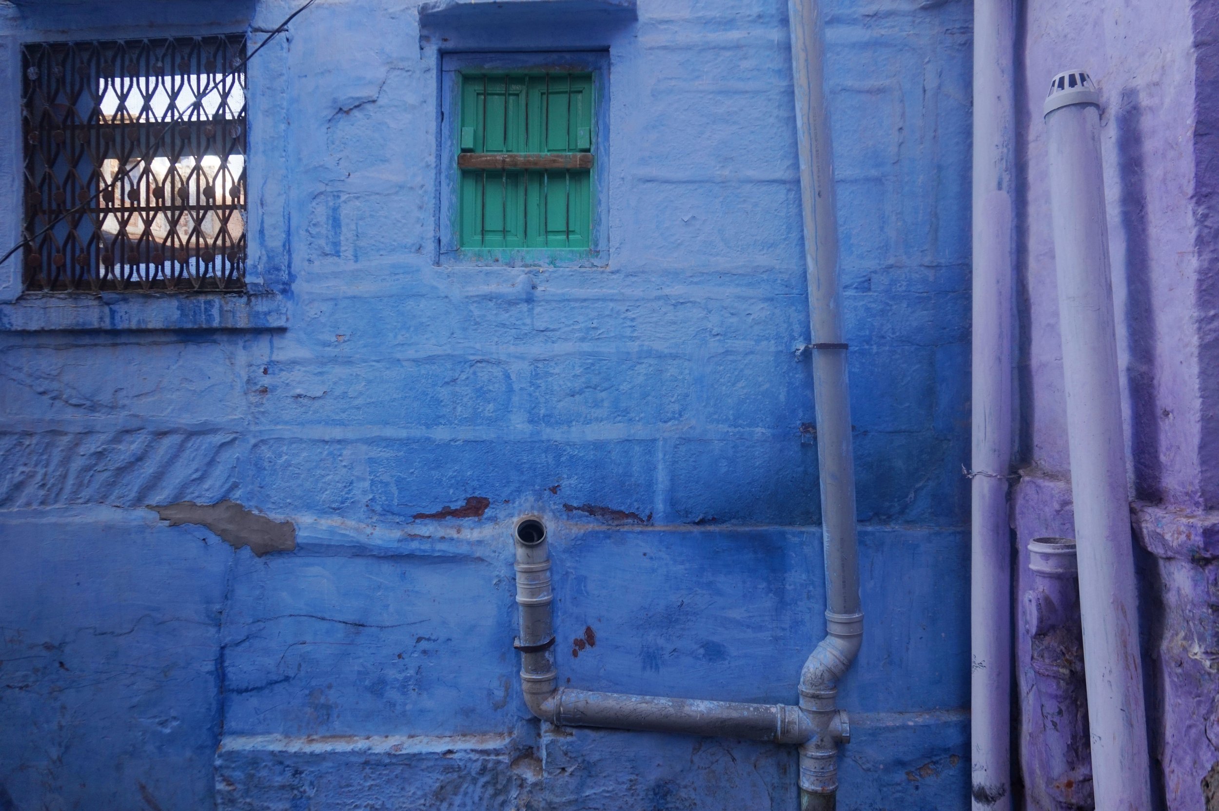 An exterior wall painted in blue with a small window with green shutters and security bars. There are white pipes along the wall, and a metal grate on the left side. The wall has visible cracks and weathering.