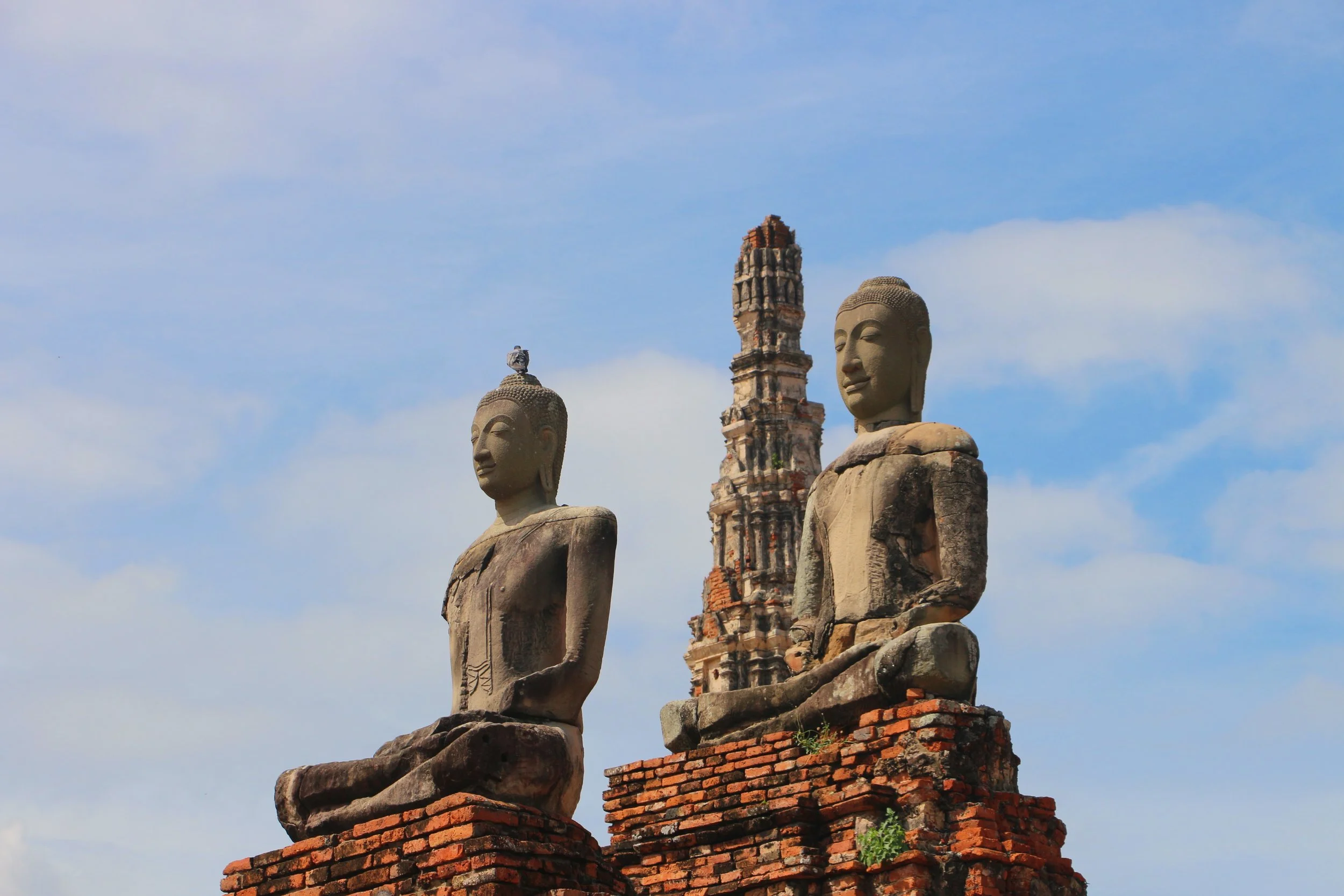 Ancient stone and brick statues of two Buddhist figures with a ruined brick pagoda in the background, set against a cloudy sky.