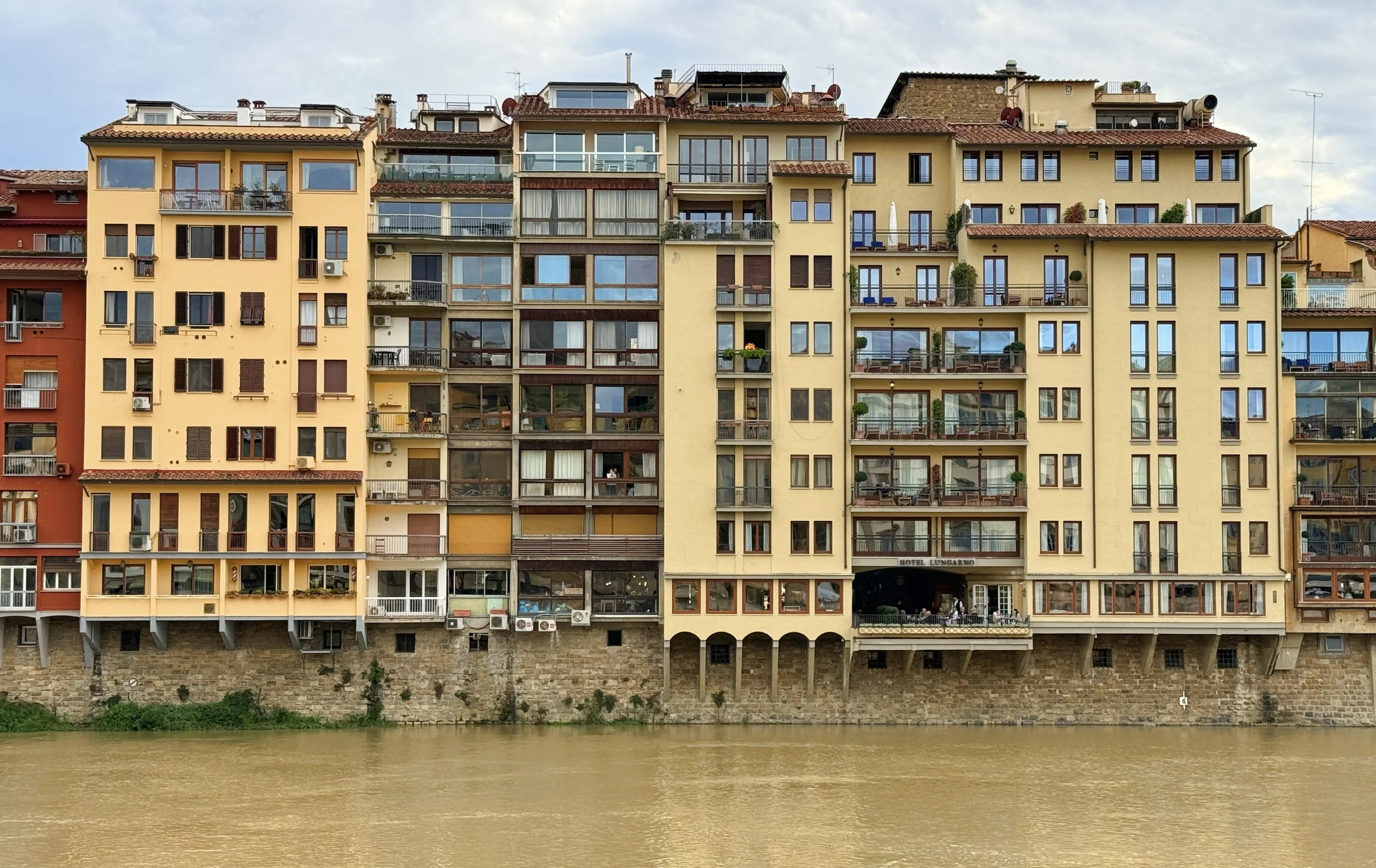 Colorful multi-story apartment building along riverbank with balconies and windows, with a hotel labeled 'Hotel Lungarno' on ground floor, under a partly cloudy sky.