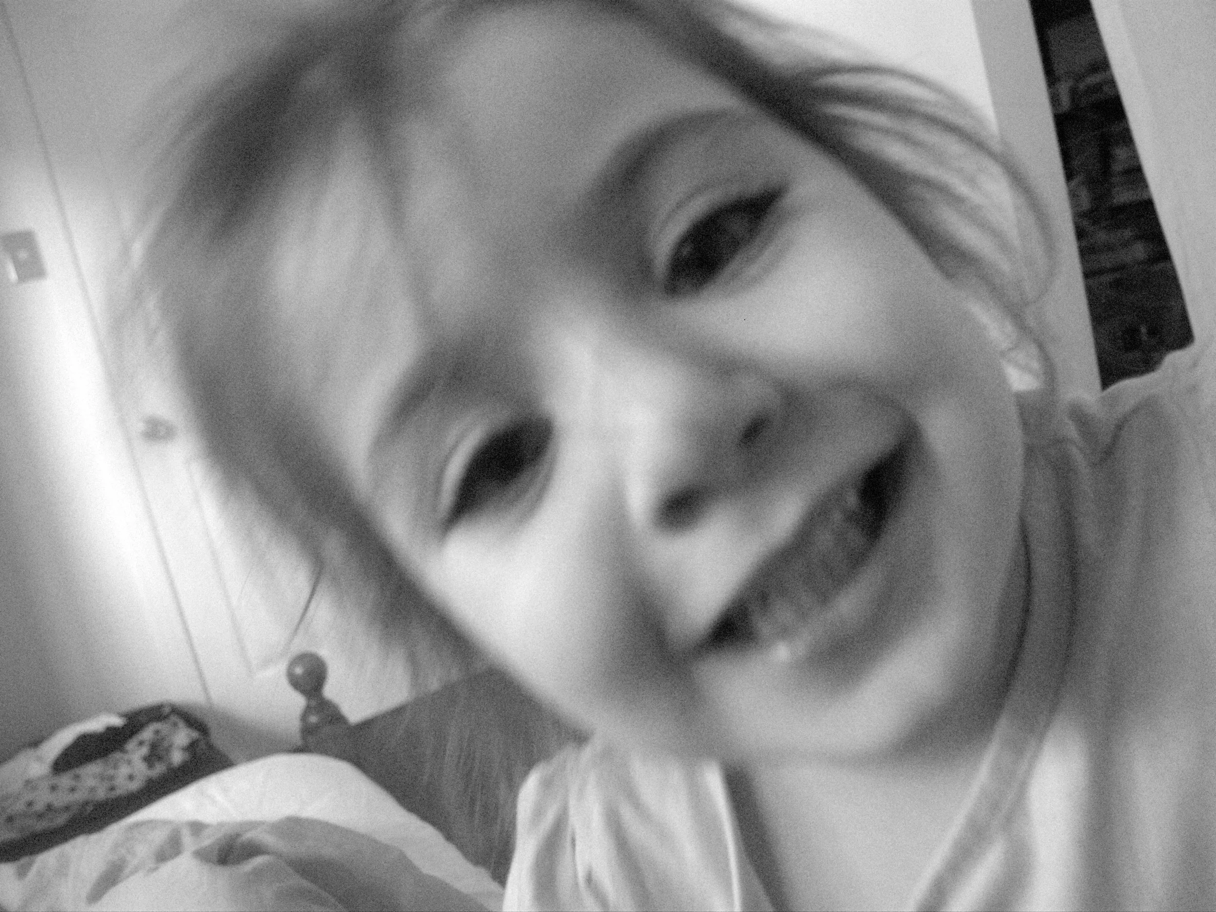 A close-up of a person's face, showing an eye and part of their forehead, in black and white. The photo appears to be taken at an angle with some background elements, including a wall and a shelf.