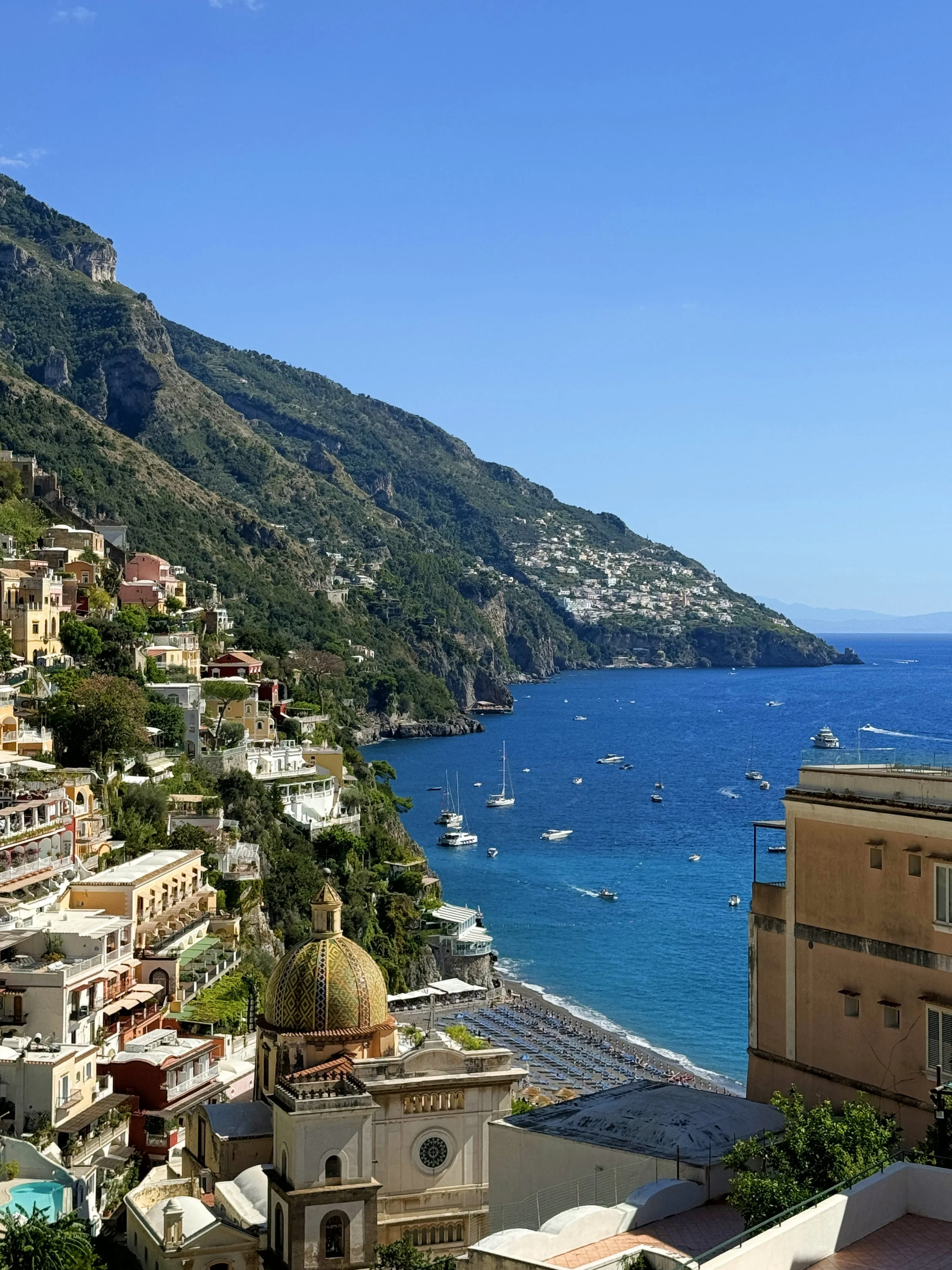 Coastal hillside with colorful buildings, a church with a domed roof, and boats in the blue sea under a clear sky.