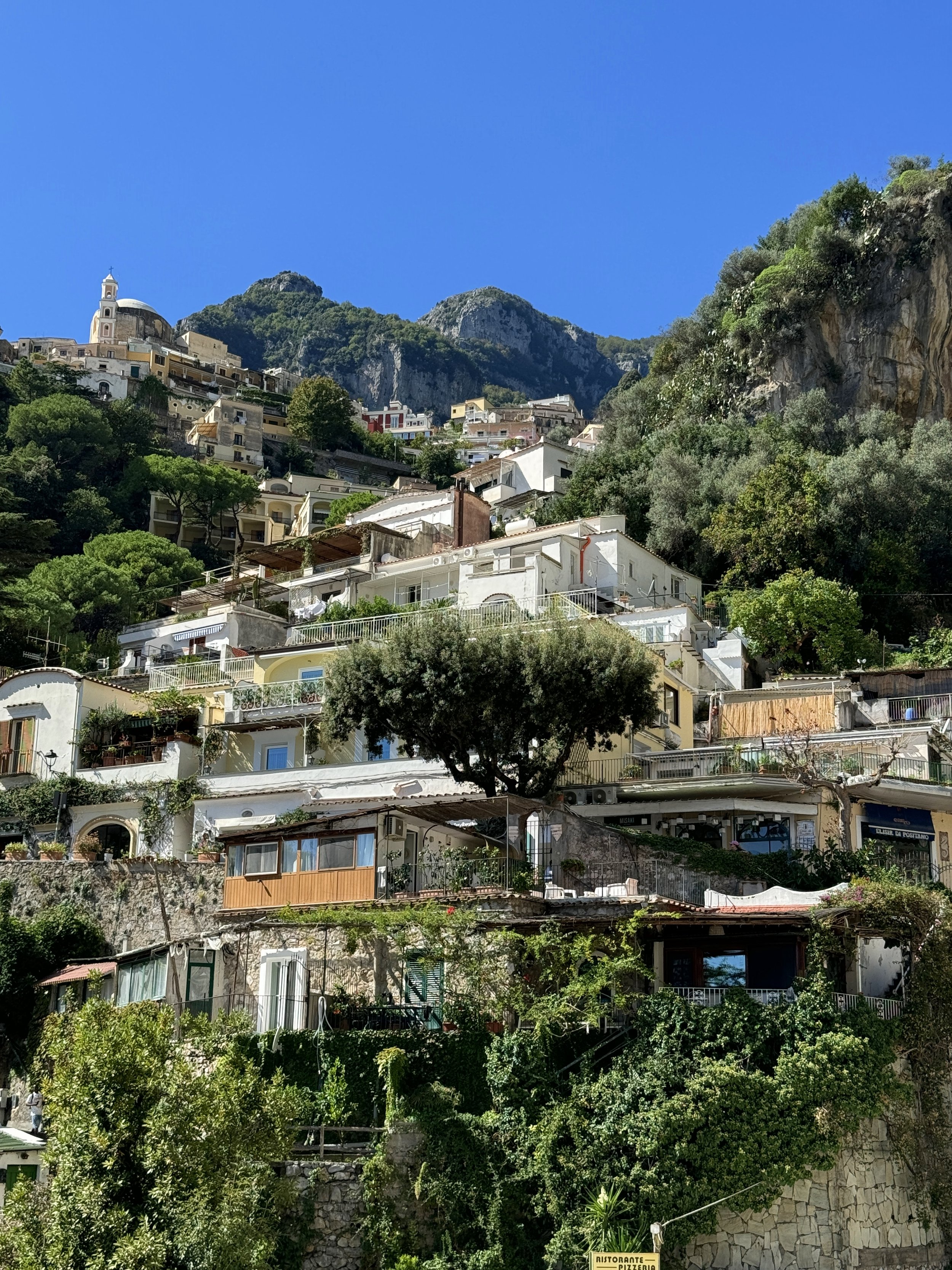 A hillside with many white and pastel-colored buildings, including a church with a dome and bell tower, surrounded by greenery and mountains under a clear blue sky.