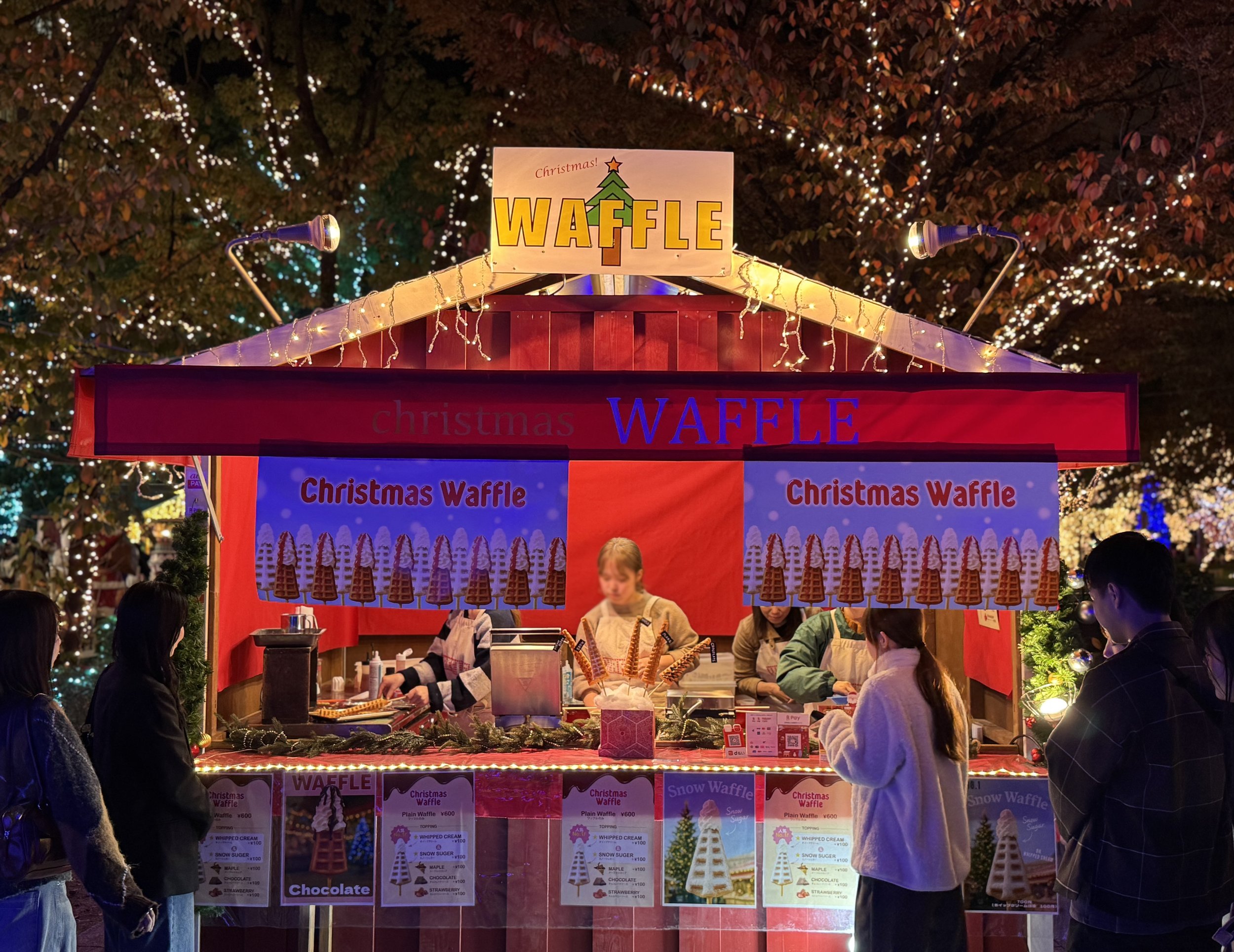 A festive Christmas market stall selling waffles at night, decorated with string lights, a sign with a Christmas tree, and surrounded by people.