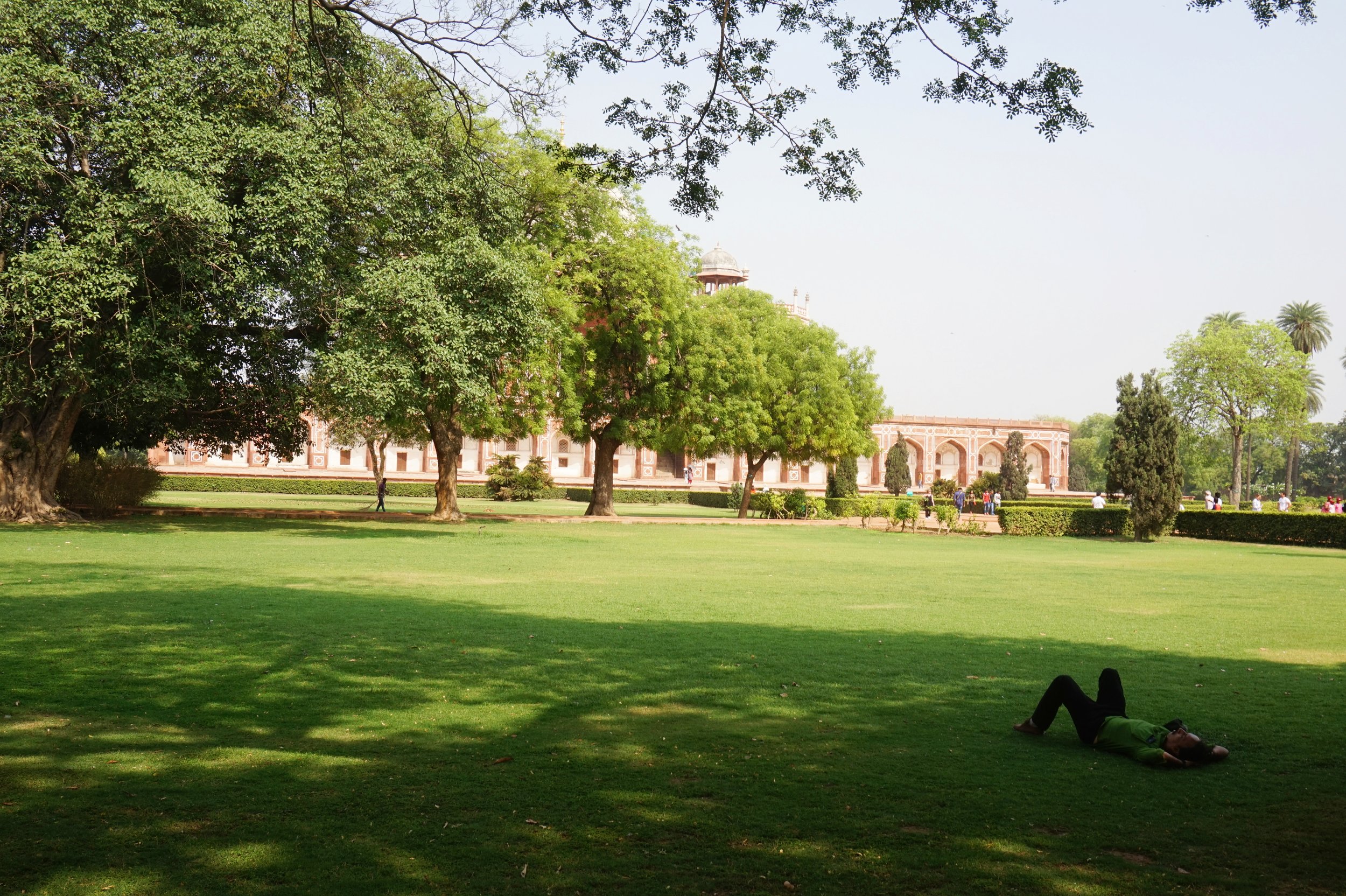 A person lying on the grass in a park with large trees, a historic building with arches and a dome, and people walking and sitting in the background.