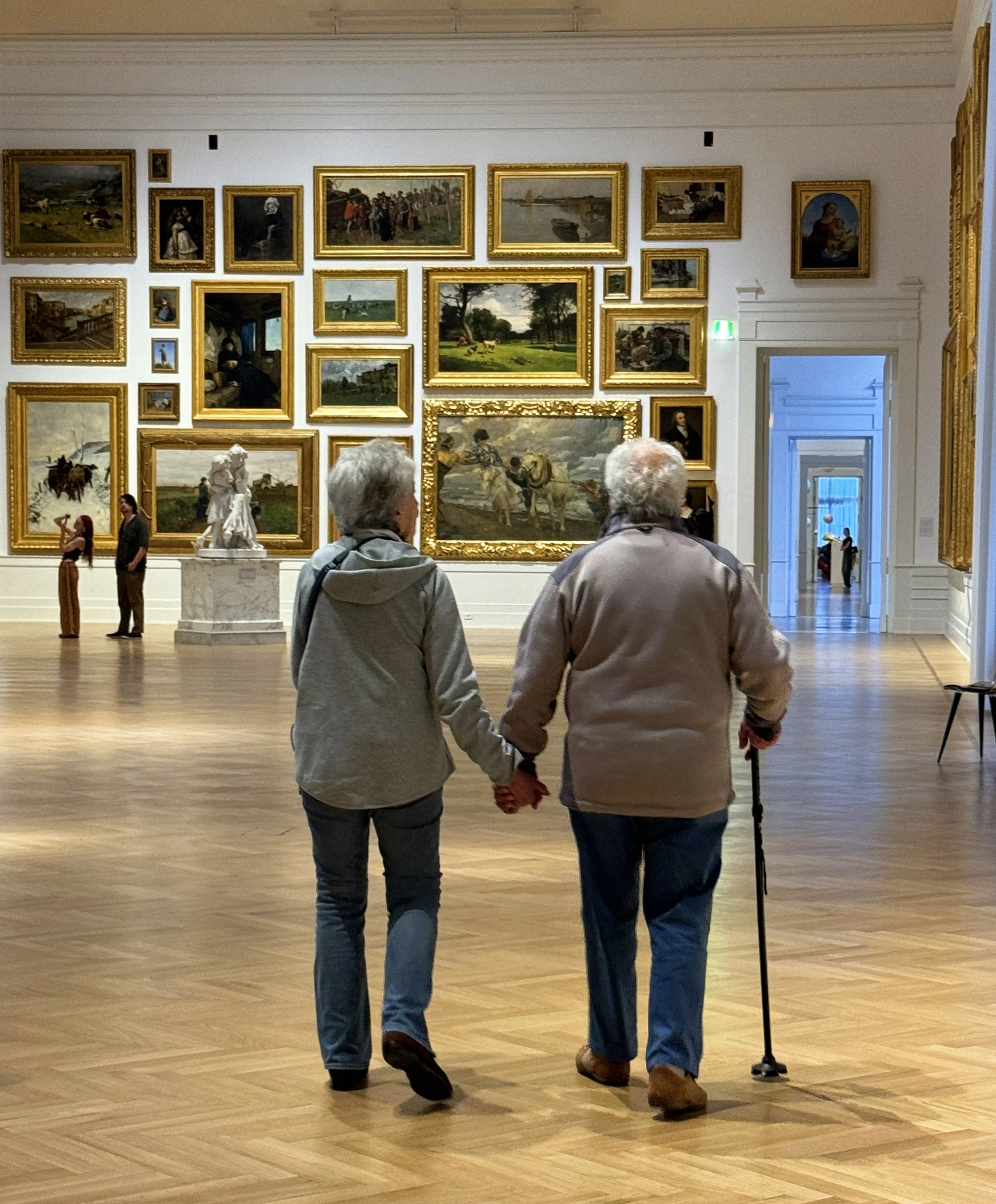 Two elderly people walking hand in hand through an art gallery, viewing paintings on the wall in a spacious room with wooden floors.