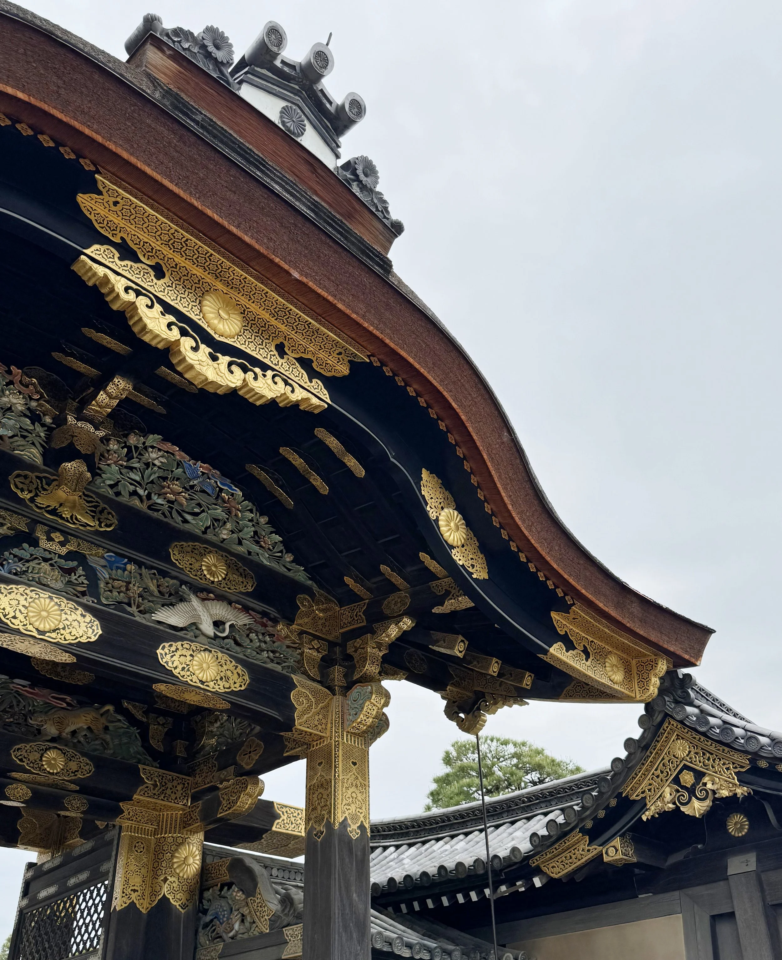 Detailed close-up image of a traditional Japanese temple roof with intricate gold and black ornamentation, ornamental tiles, and decorative elements, set against a cloudy sky background.