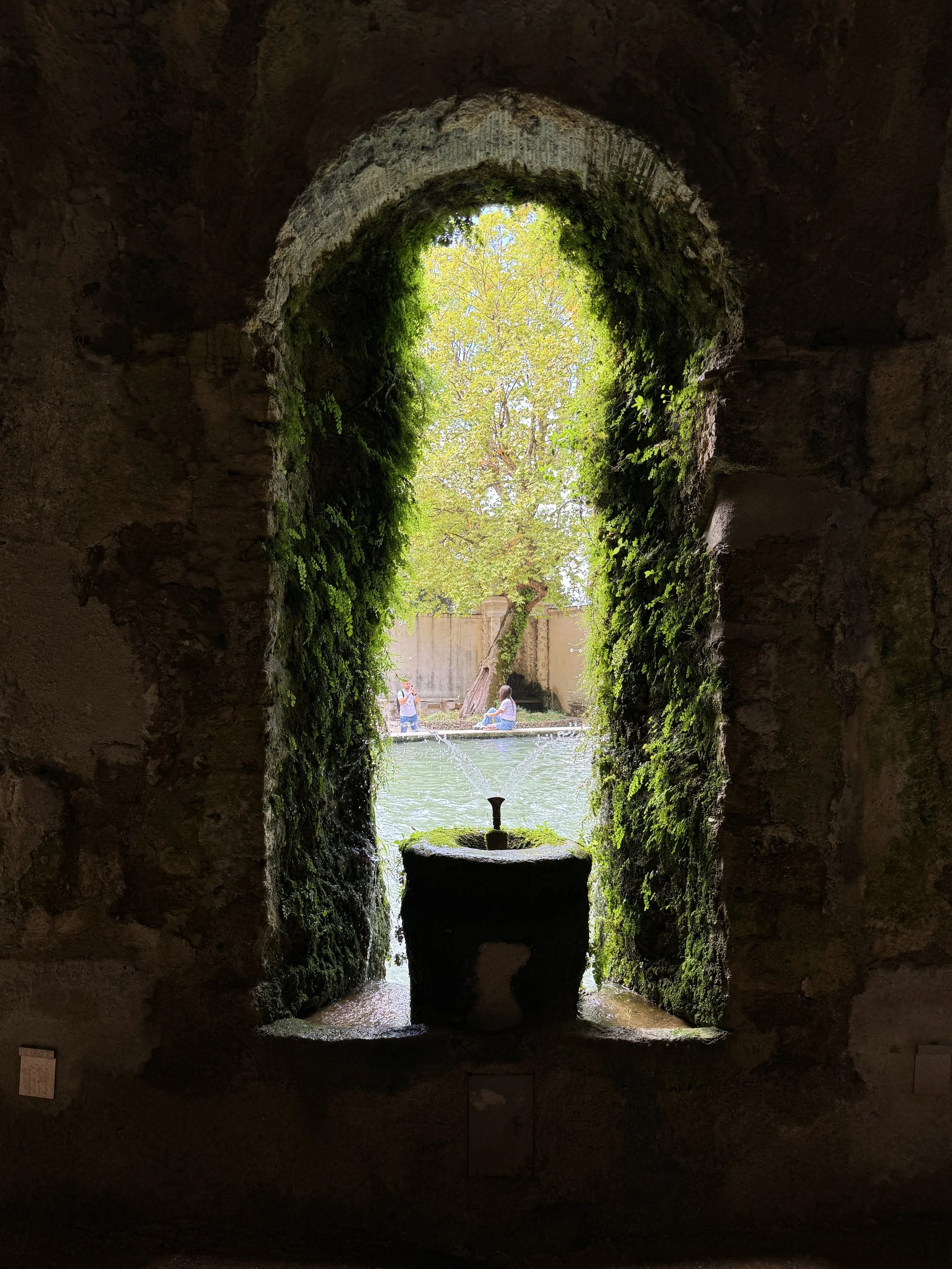 View from inside a stone archway looking out to a pond with two people sitting on the edge, surrounded by trees and greenery.