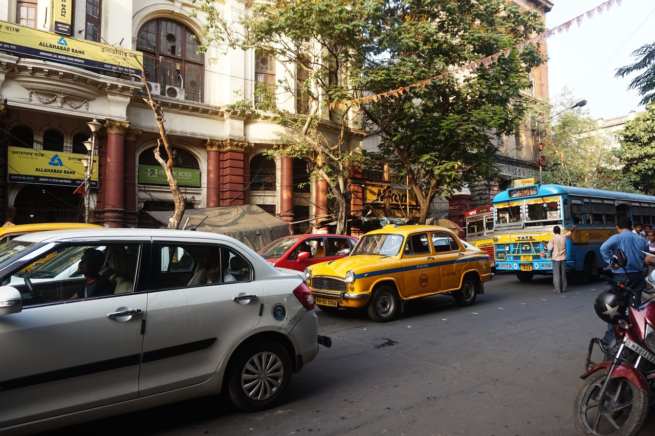 Street scene with various vehicles, including a silver car, a yellow taxi, and a blue bus, in front of a building with trees and banners hanging overhead.