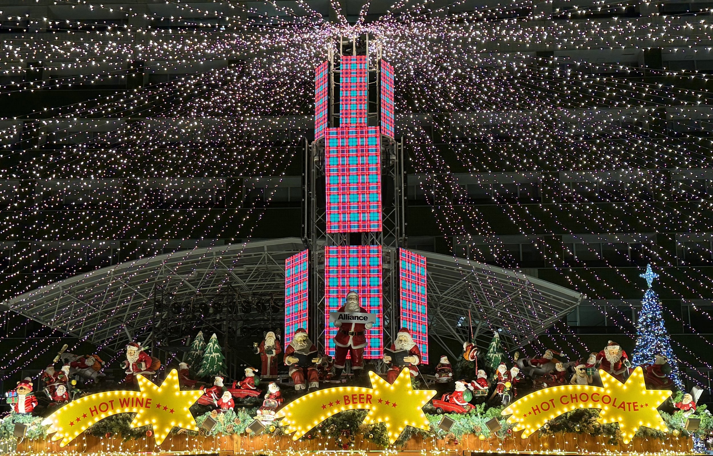 Christmas market stall decorated with lights and festive figures including Santa Claus, with signs advertising hot wine, beer, hot chocolate, and other treats.