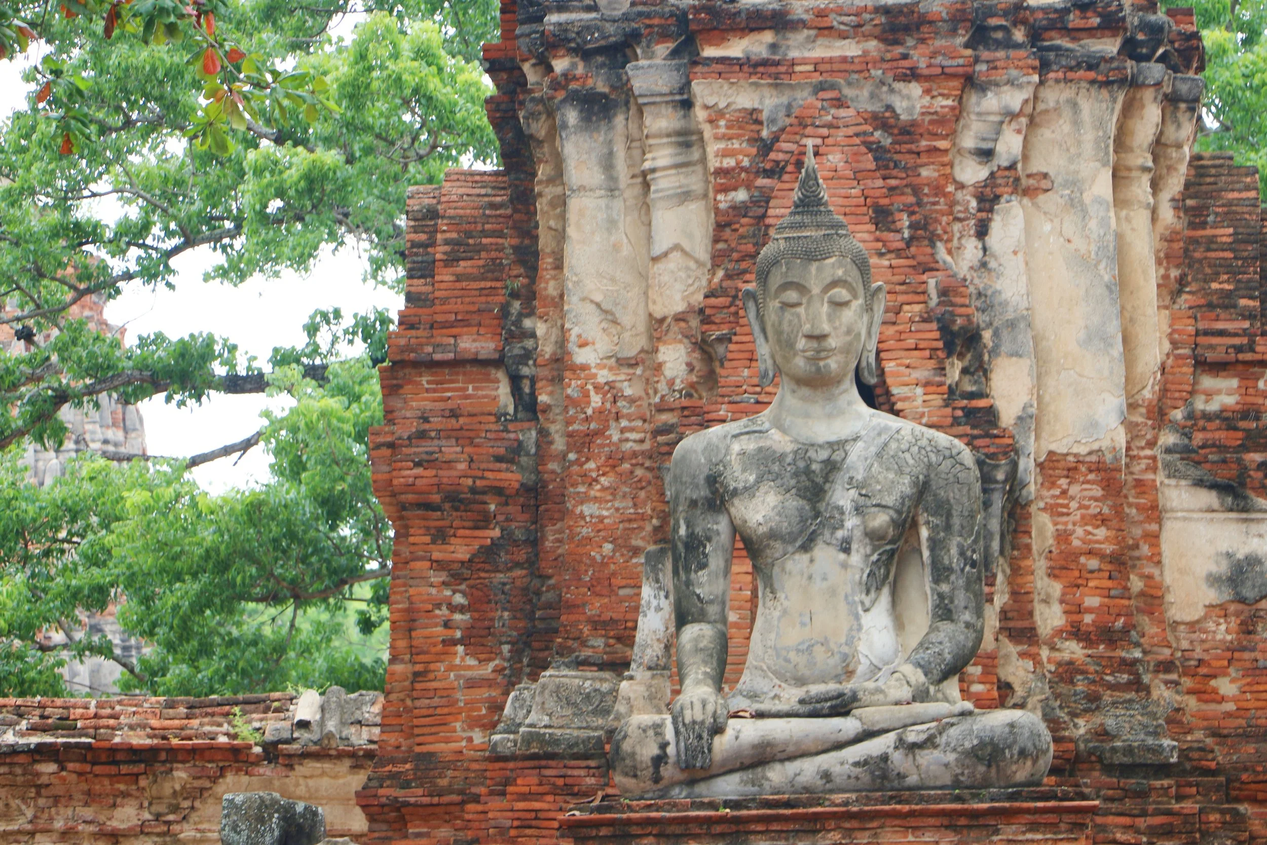 Ancient stone Buddha statue in front of a weathered brick building, surrounded by green leafy trees.