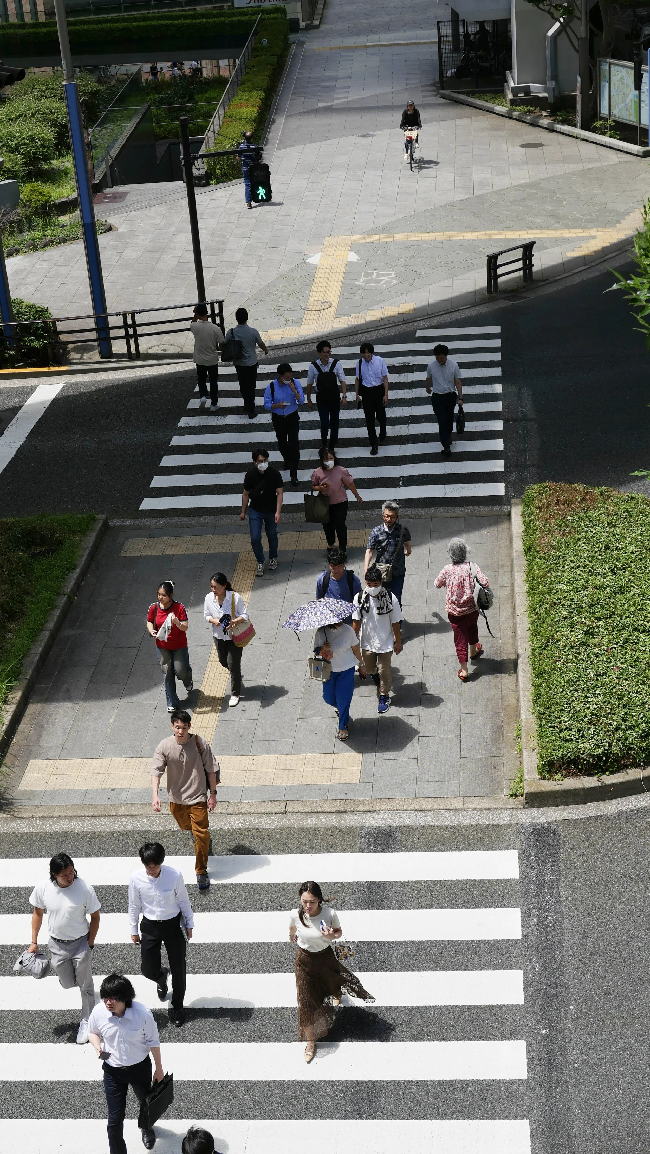 People crossing a crosswalk on a city street during daytime, some wearing masks, with a sidewalk, road, and greenery in the background.