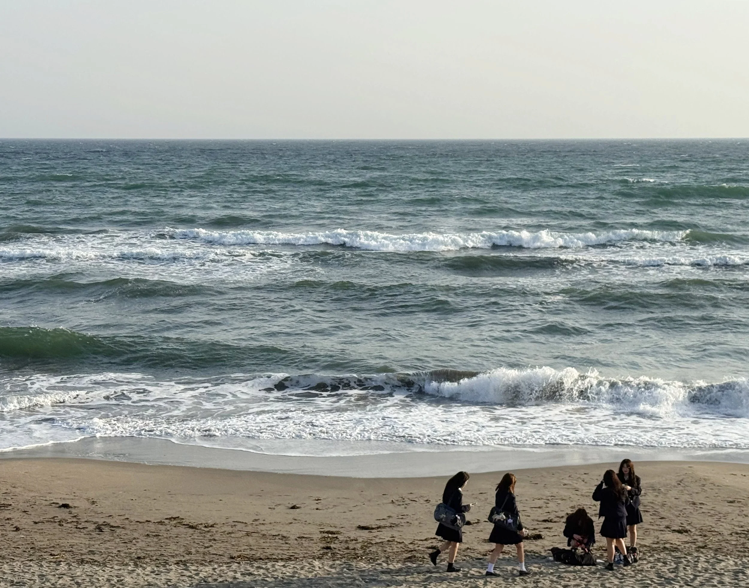A beach scene with four people walking and talking on the sand near the shoreline, with waves and the ocean in the background.