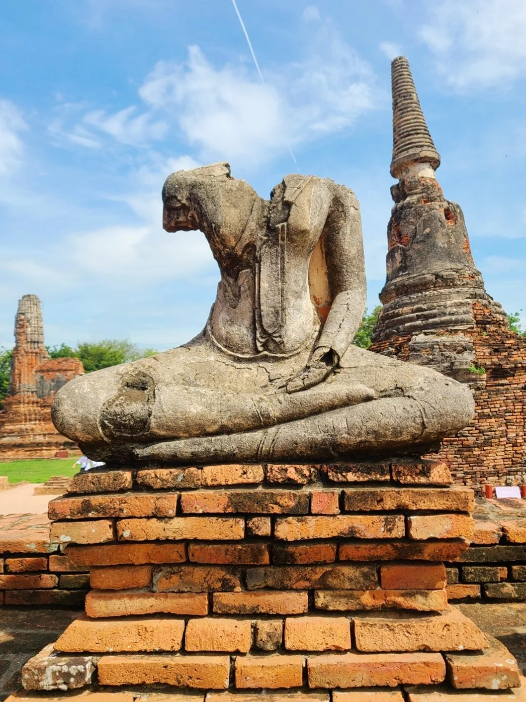 Ancient stone Buddha statue sitting cross-legged with missing head, located in a historical site with brick stupas and ruins in the background under a blue sky.