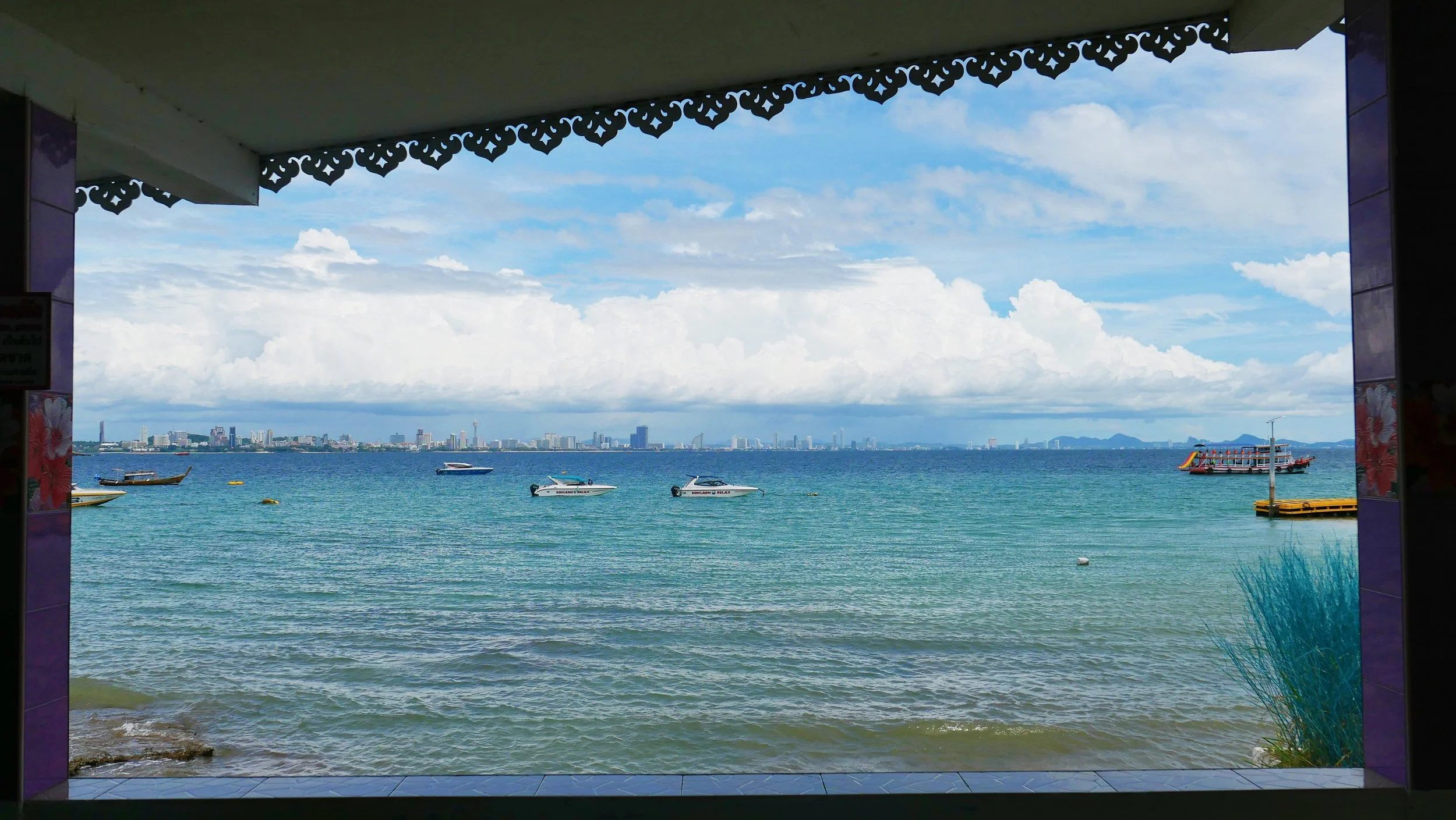 View of a city skyline across the water, seen from an opening framed by ornate decorative trim overhead, with boats and a ferry on the water and a cloudy sky.