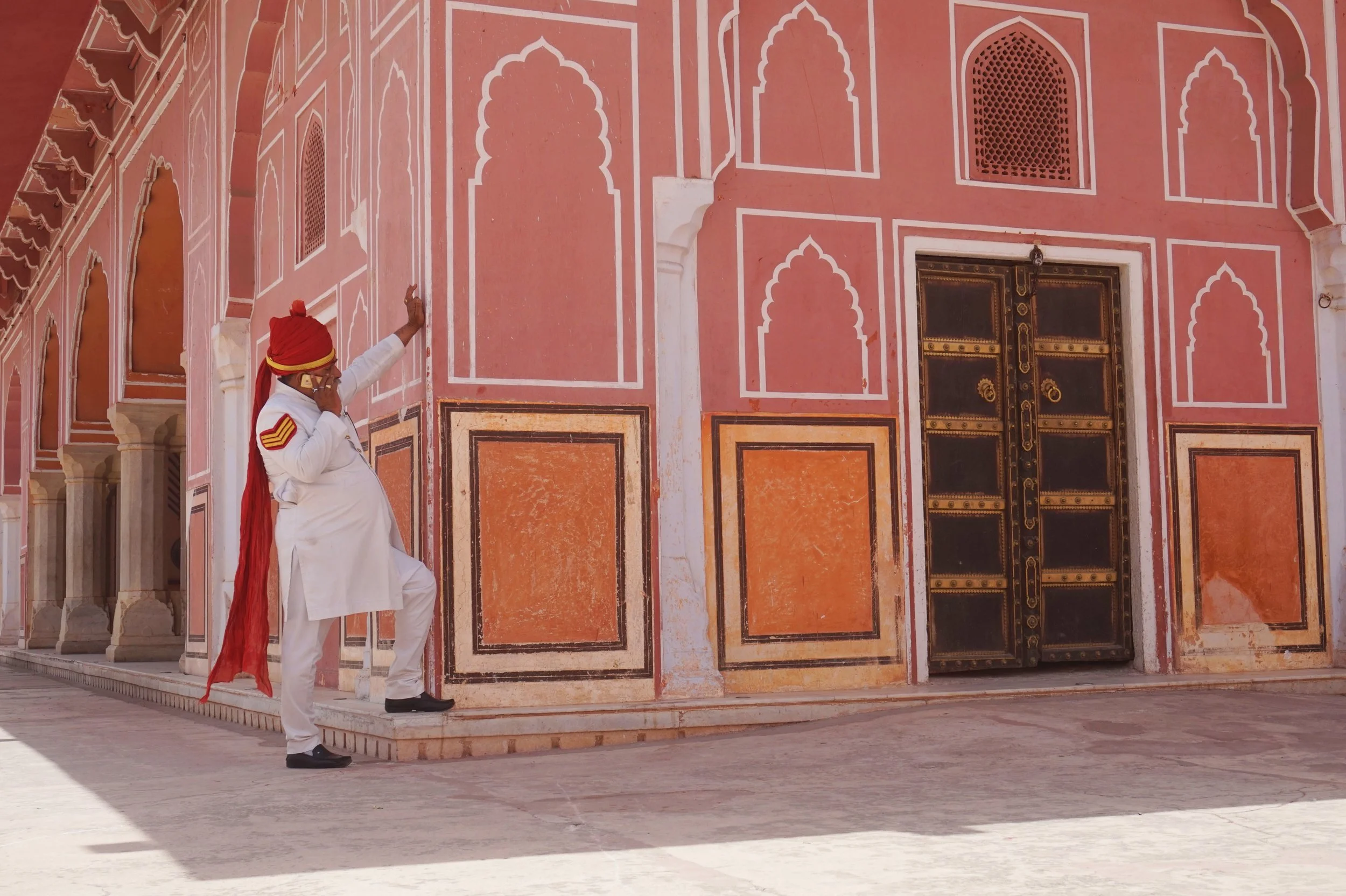 A man dressed in traditional Indian wedding attire, including a white kurta, black shoes, and a red turban, is standing and touching the wall of a pink and orange palace with intricate white and black designs. The palace wall has arched windows and a