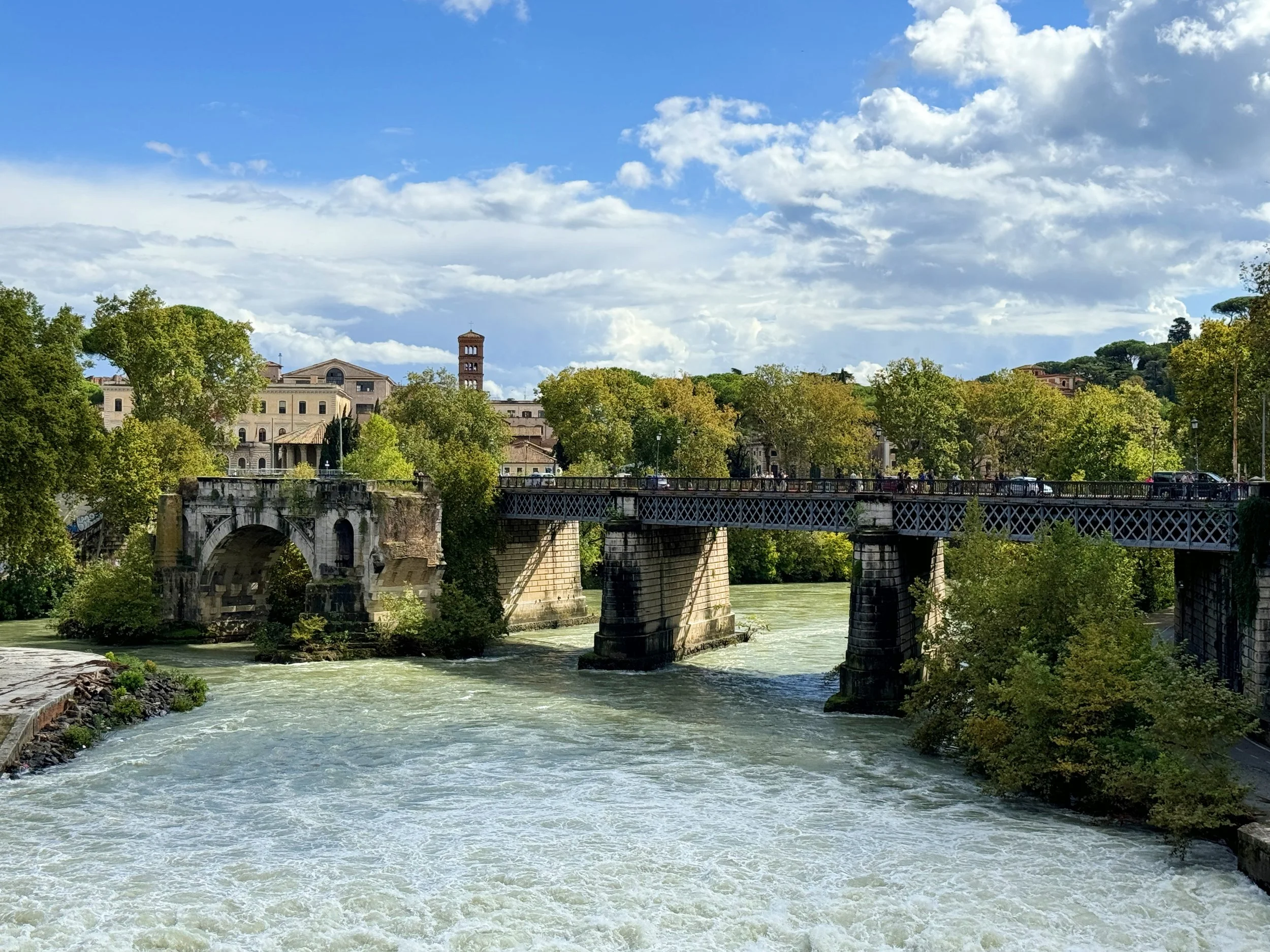 A river flowing under an old stone bridge, with trees and historic buildings in the background, under a partly cloudy sky.