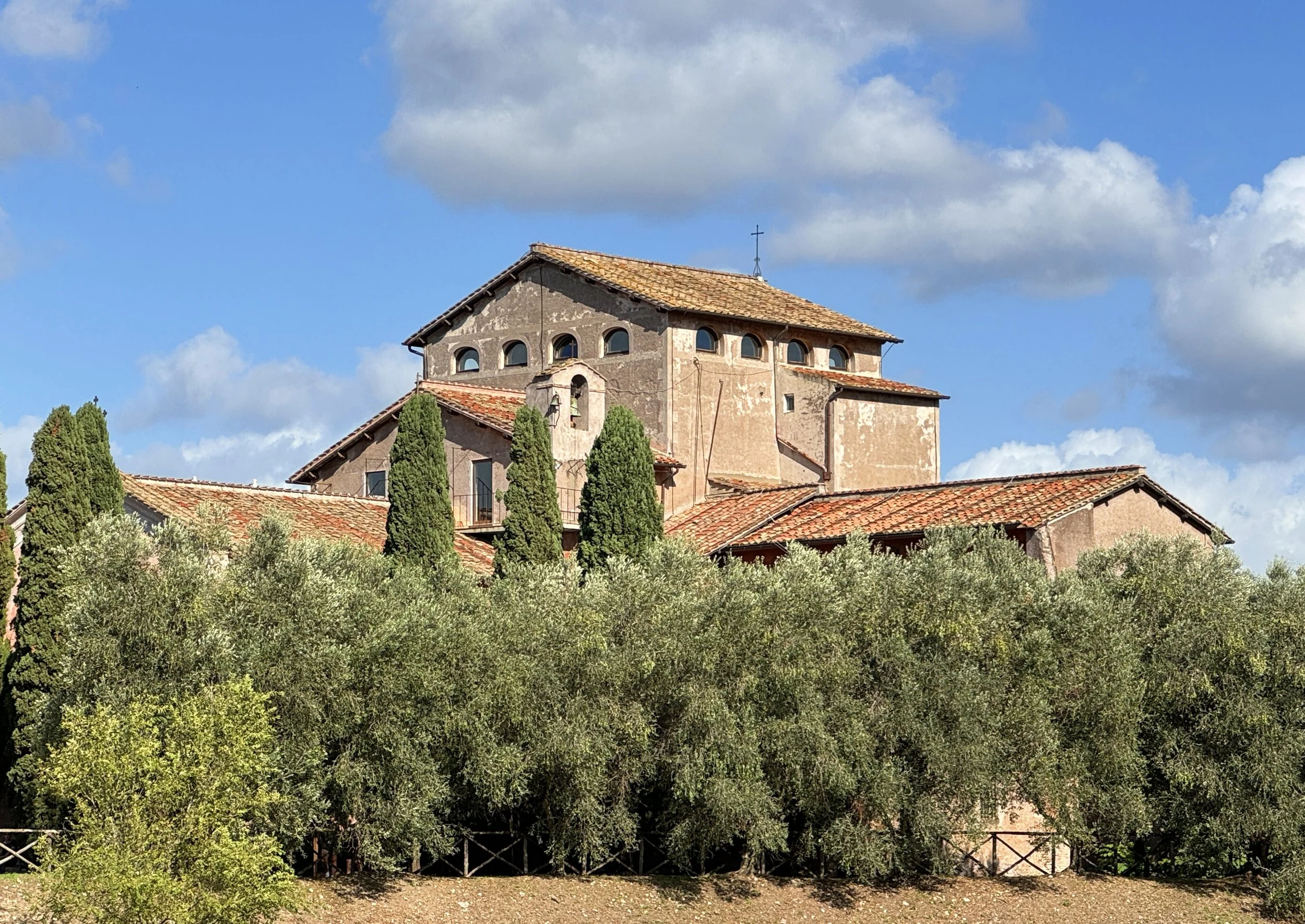 A rustic church with a tiled roof and cross on top, surrounded by tall green trees and bushes under a blue sky with scattered clouds.