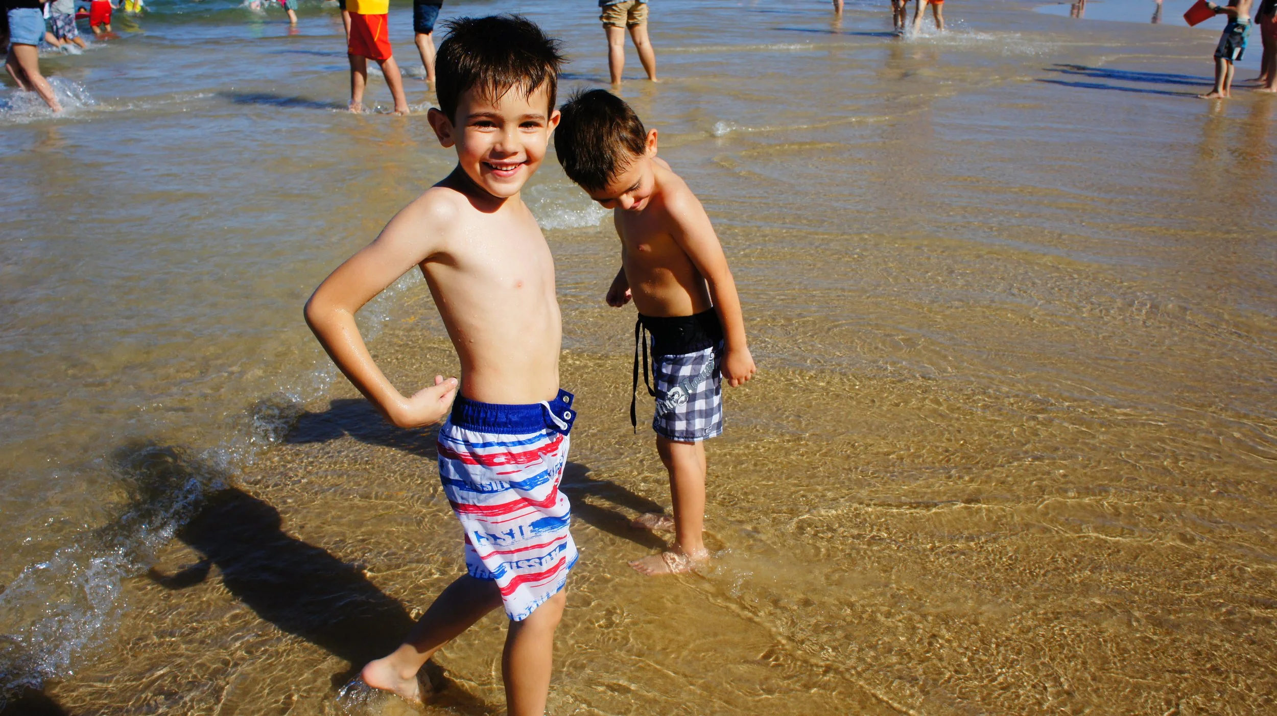 Two young boys playing in shallow water at the beach, smiling and enjoying their time with other people in the background.