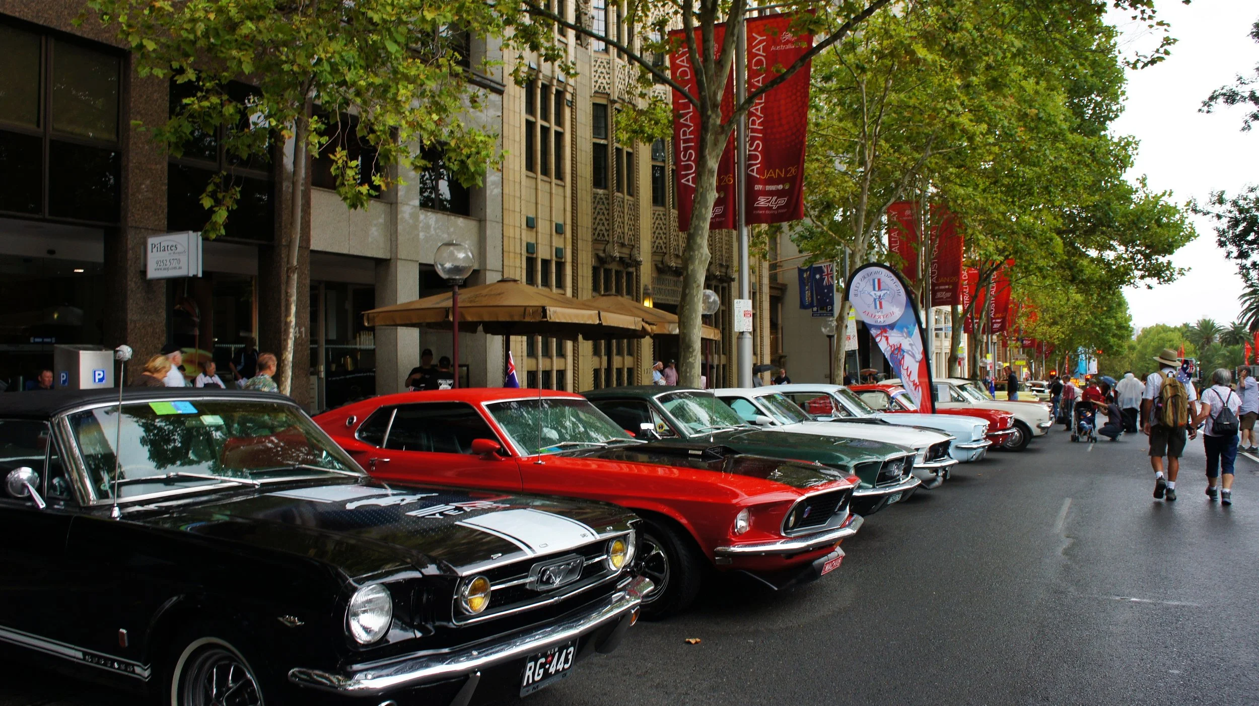 A street scene with classic cars parked along the curb and people walking. There are red banners with white text hanging from trees, and a building with ornate architecture in the background. The street is lined with trees and there are umbrellas ove