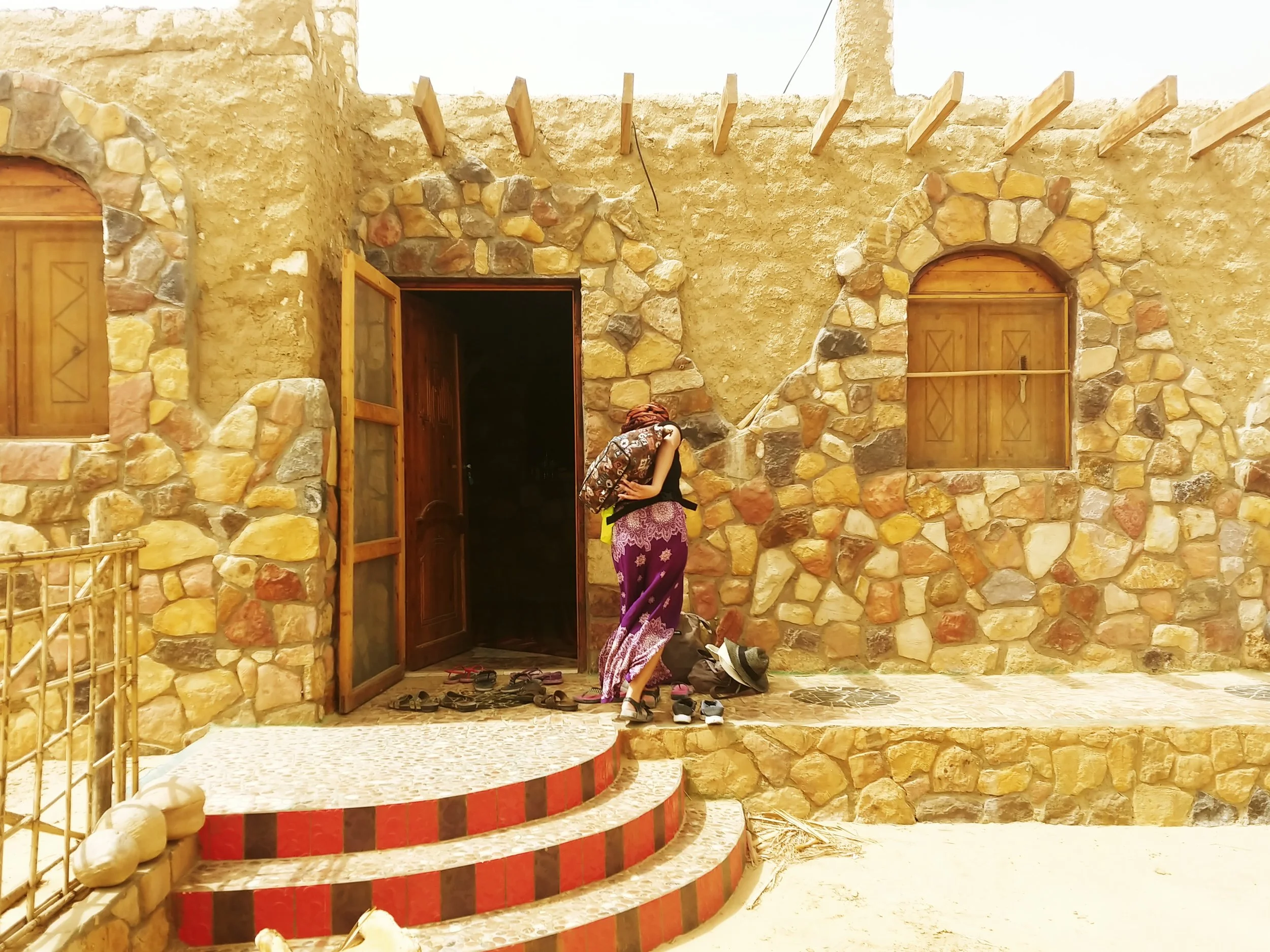 A person wearing purple patterned pants and a black shirt, is bending down, sitting on the stairs outside a stone building. They are surrounded by multiple pairs of shoes and sandals. The building has a stone exterior with two arched wooden doors and