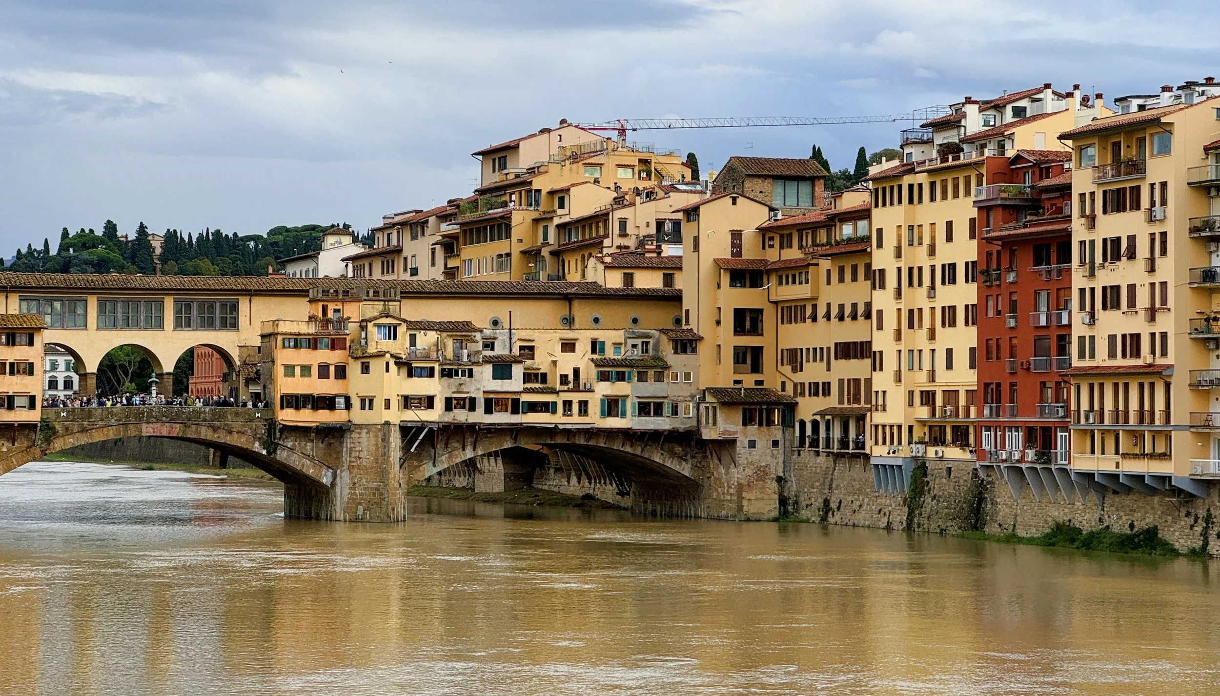 Colorful buildings in Florence, Italy, along the river with a stone bridge and a cloudy sky.
