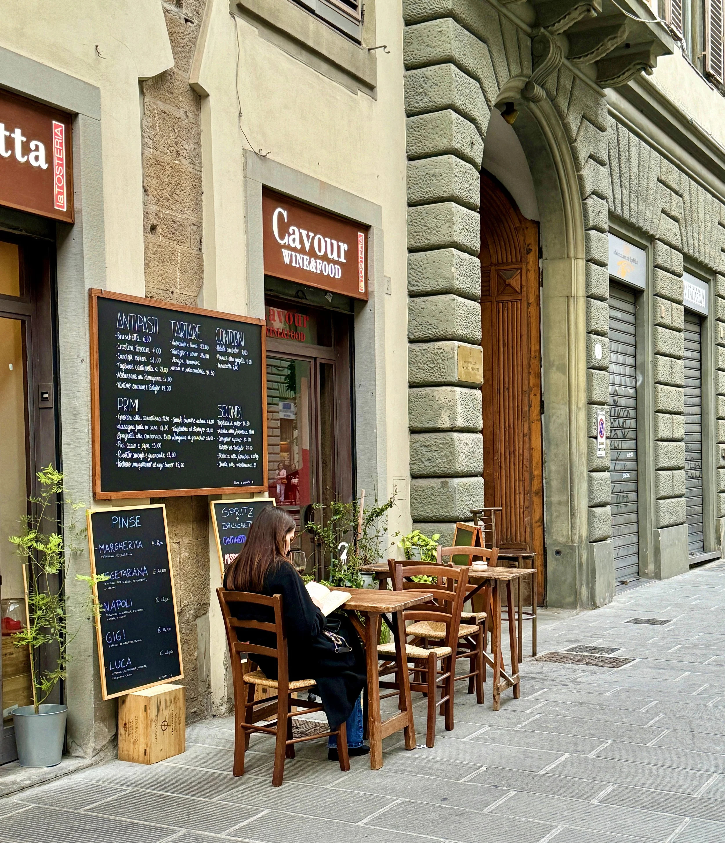 A woman with long dark hair sitting alone at an outdoor café table, reading a book. Behind her are several wooden chairs, a blackboard menu with Italian dishes, and a street view with a building featuring a stone and stucco façade. The café has signs