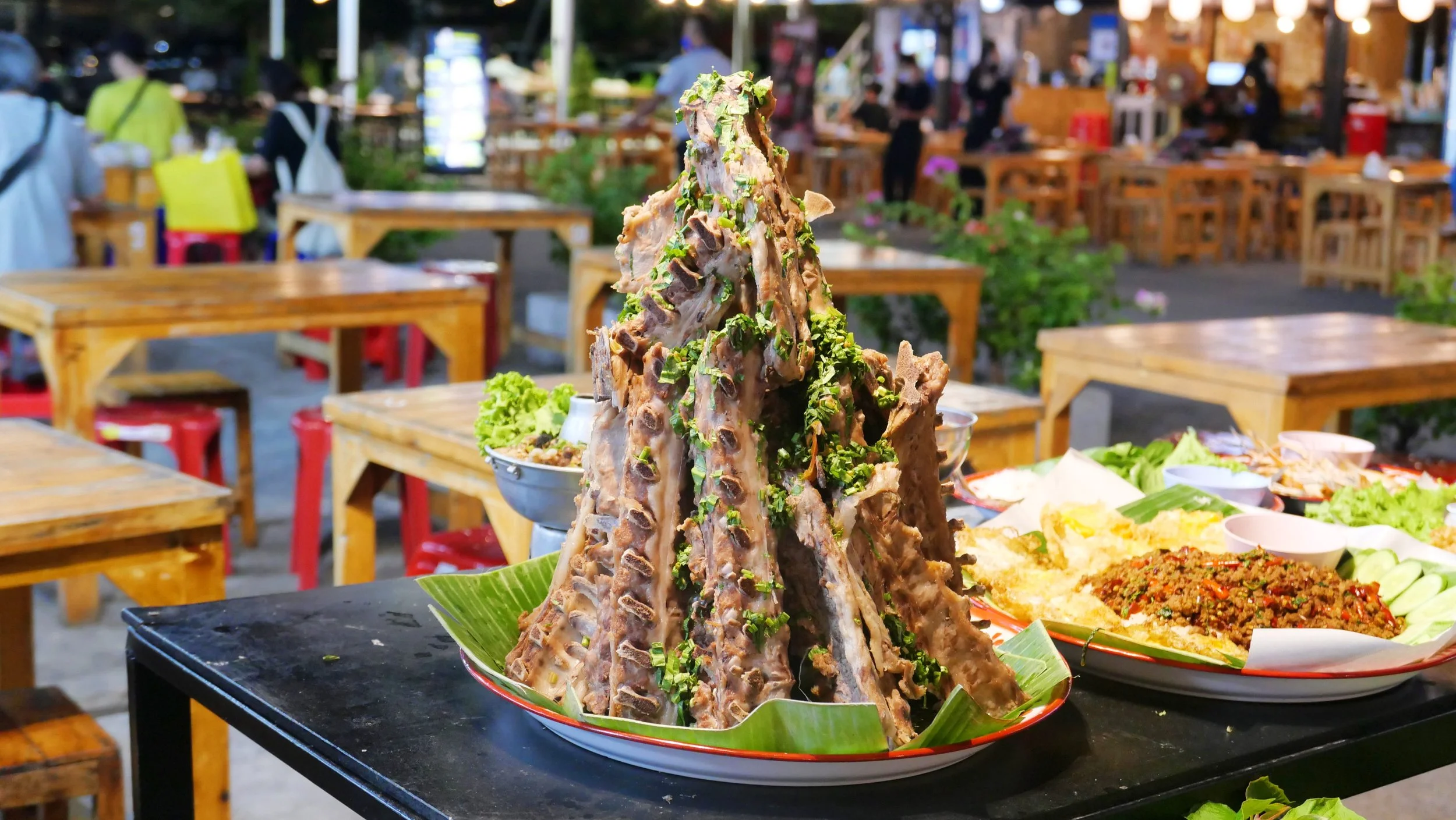 A traditional Asian dish featuring a large, bone-in meat piece, garnished with chopped herbs, set on a plate with banana leaves, in a bustling restaurant or food market setting.