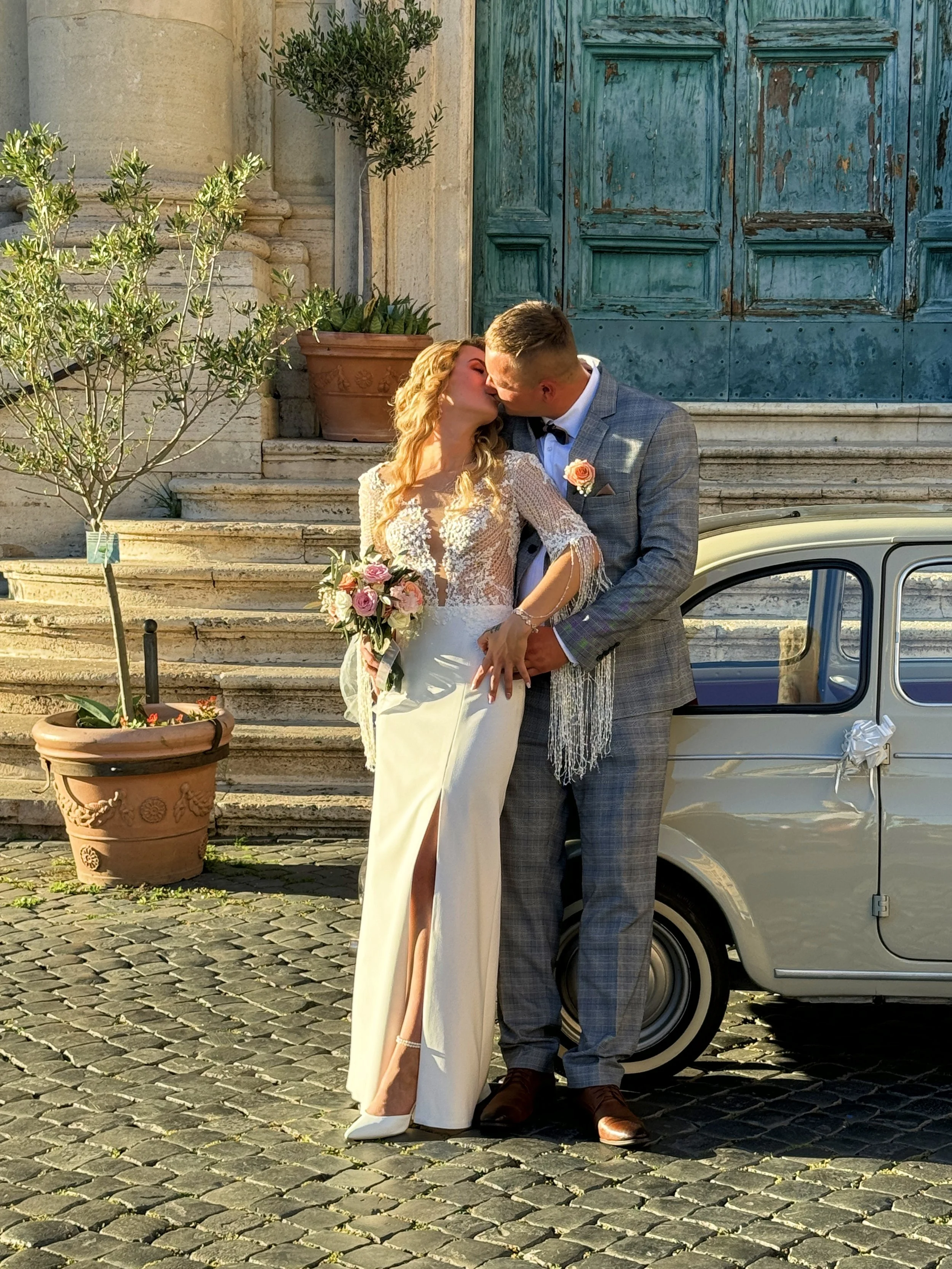 A bride and groom sharing a kiss outdoors on a cobblestone street, with potted plants, stone stairs, a large green door, and a vintage car decorated with a bow in the background.