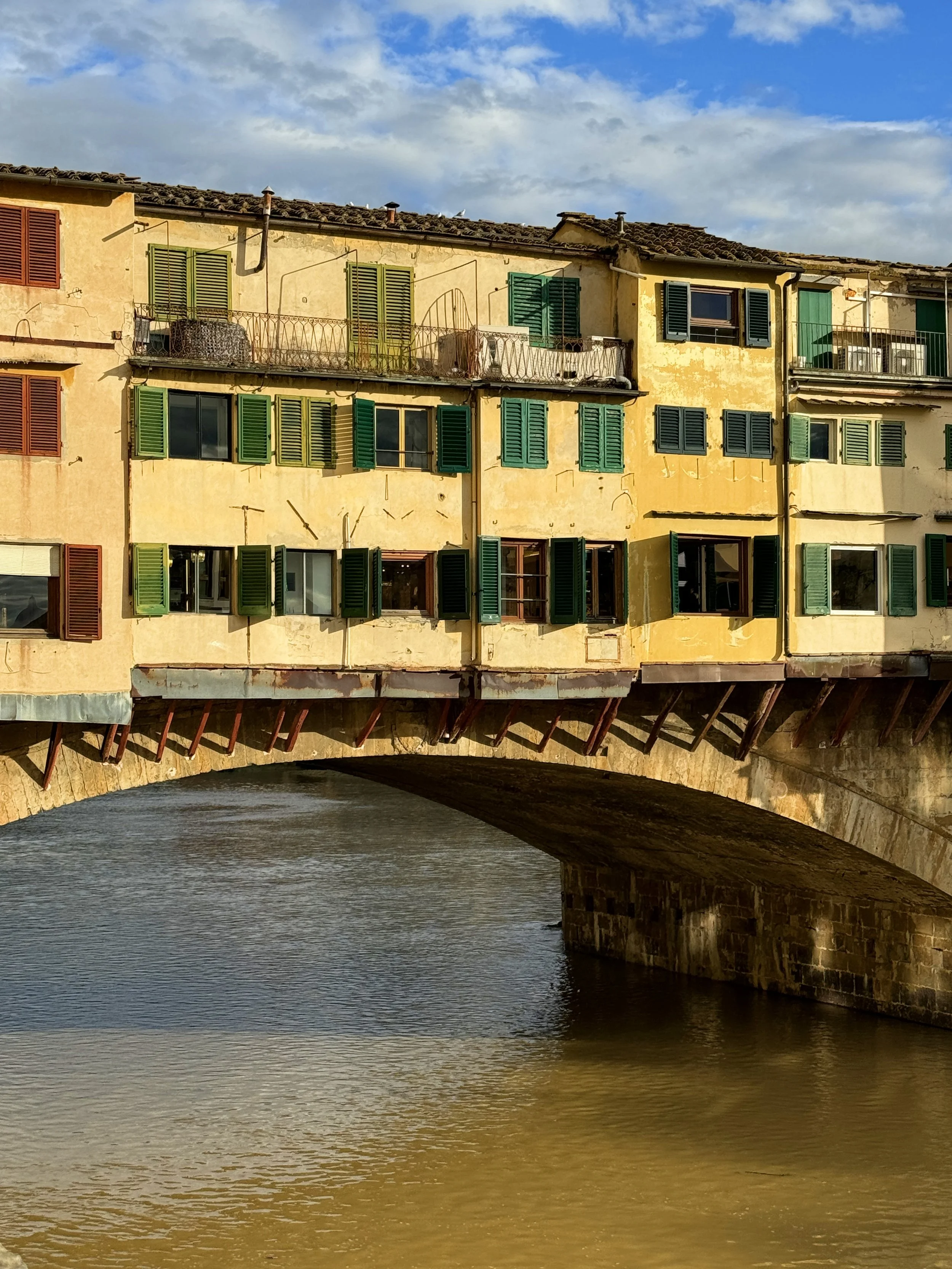 A bridge over a river with colorful buildings with green and red shutters, some with balconies, under a cloudy blue sky.