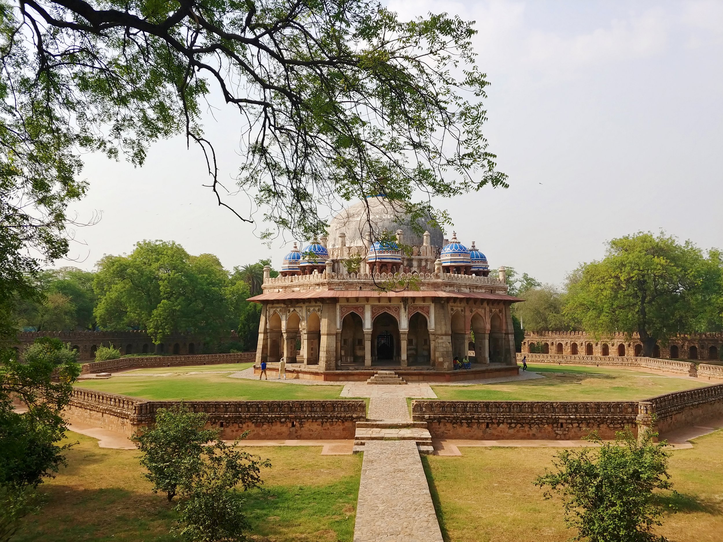 A historical monument with blue domes, ornate architecture, and an arched entrance, set within a lush green park with trees and walkways.