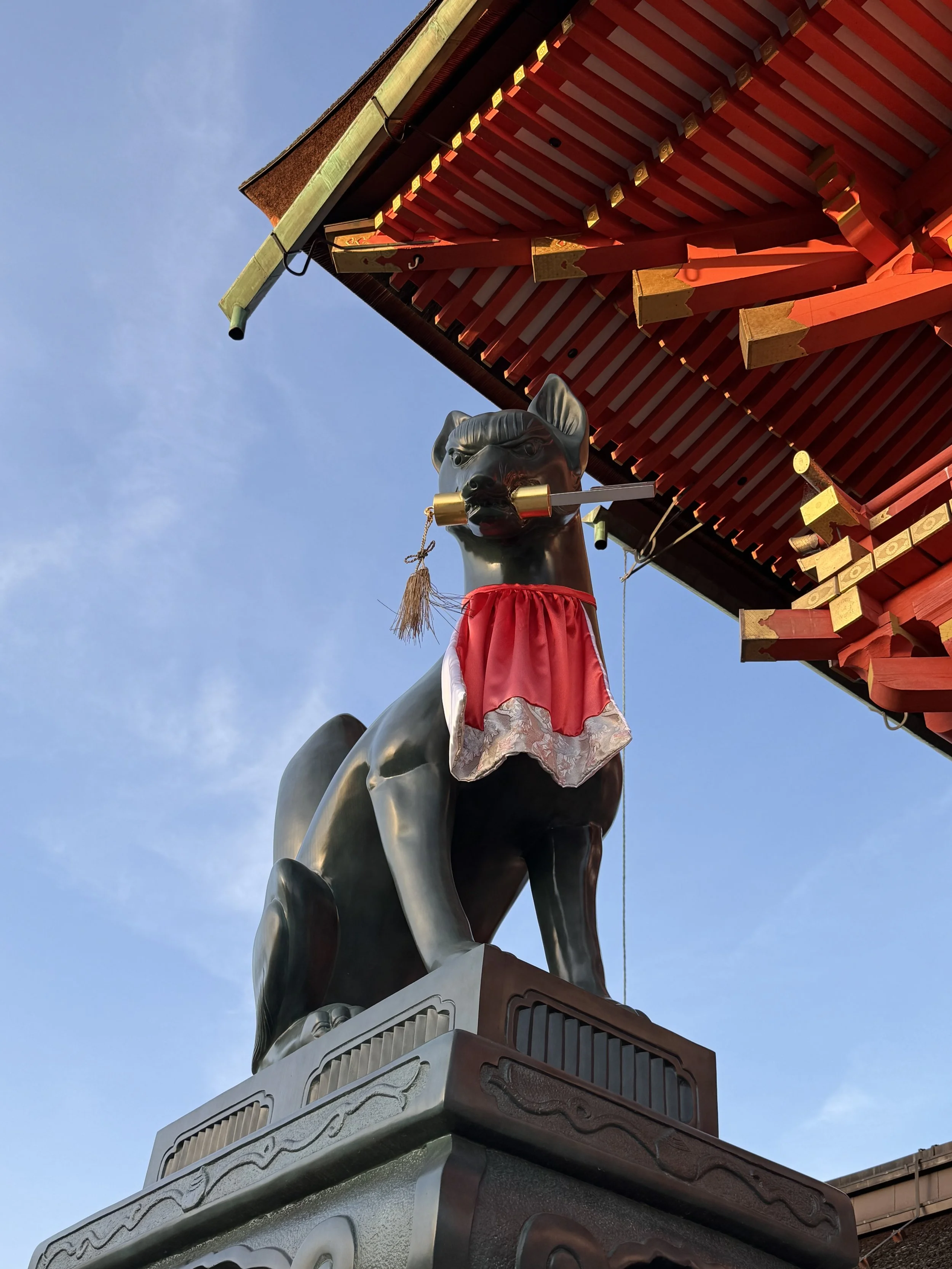 A statue of a dog with a sword in its mouth, dressed in traditional Japanese armor, located beneath the corner of a red and gold ornate roof structure against a clear blue sky.