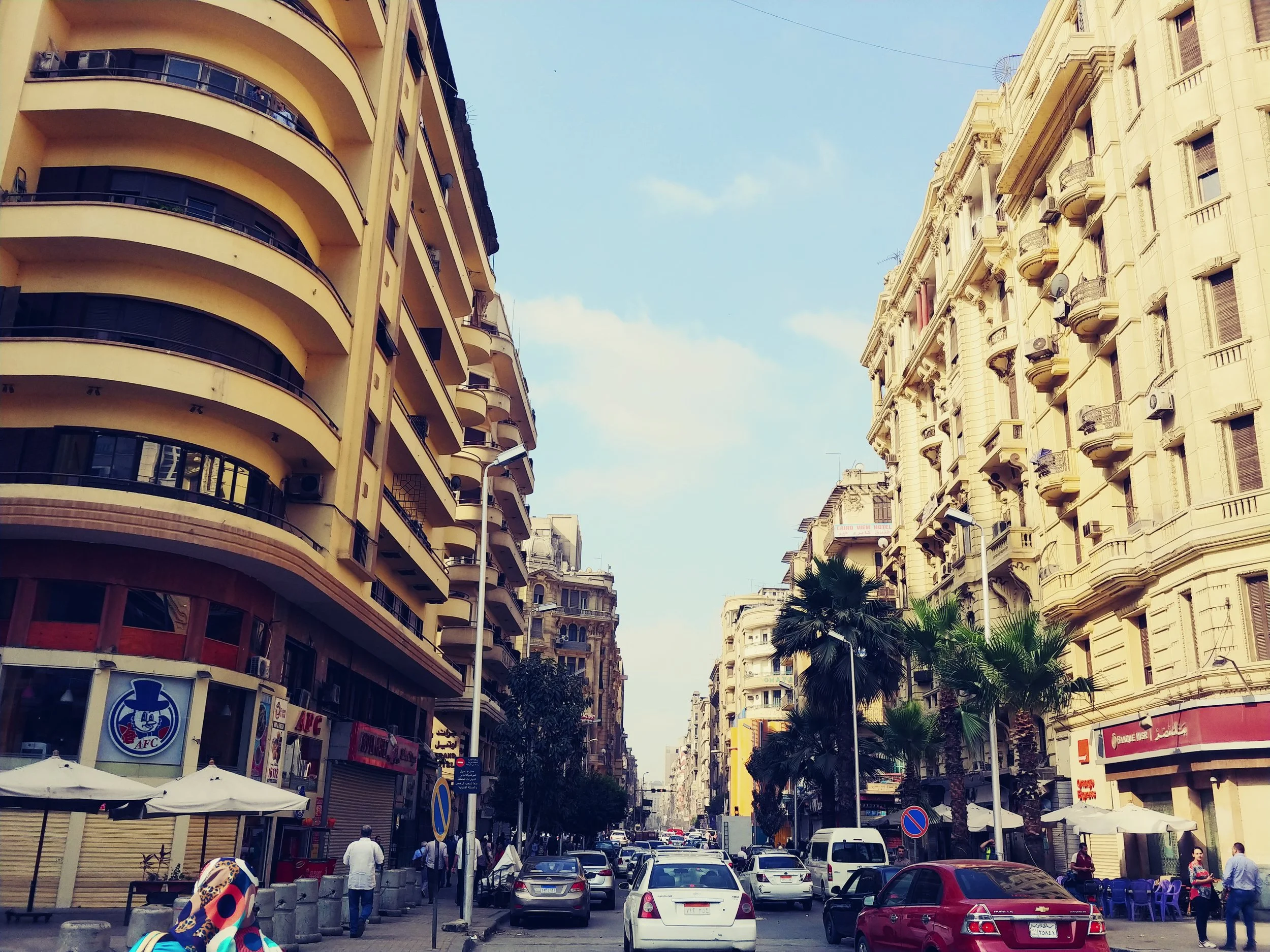 A city street with tall buildings on both sides, street cars, palm trees, and people walking.
