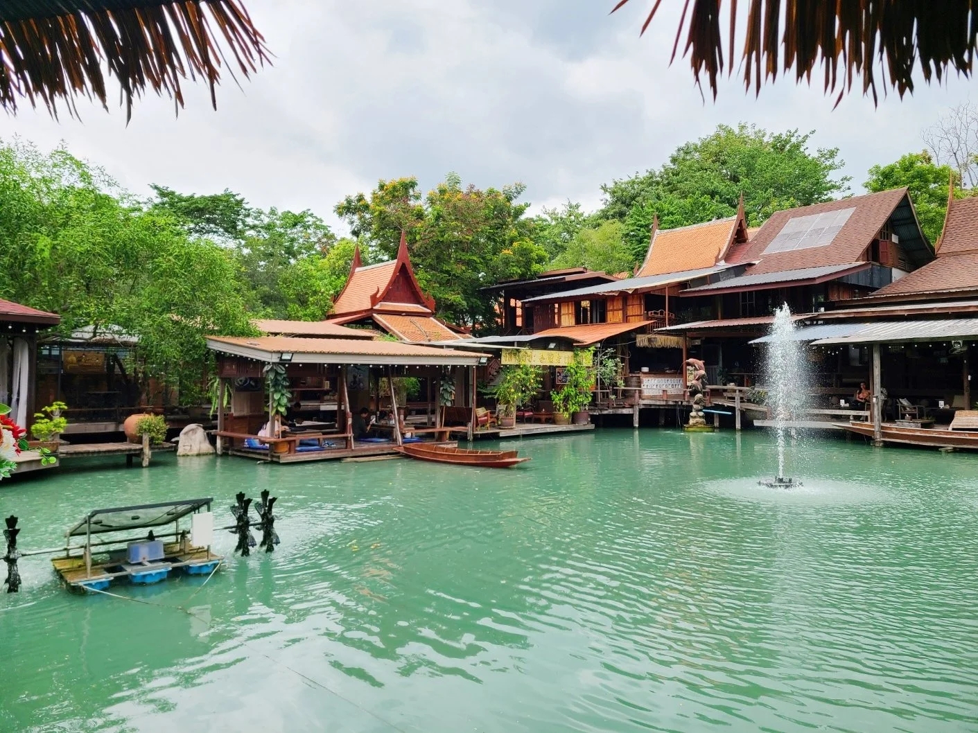 Traditional Thai-style buildings with sloped roofs surrounding a turquoise pond with a fountain, greenery, and a cloudy sky