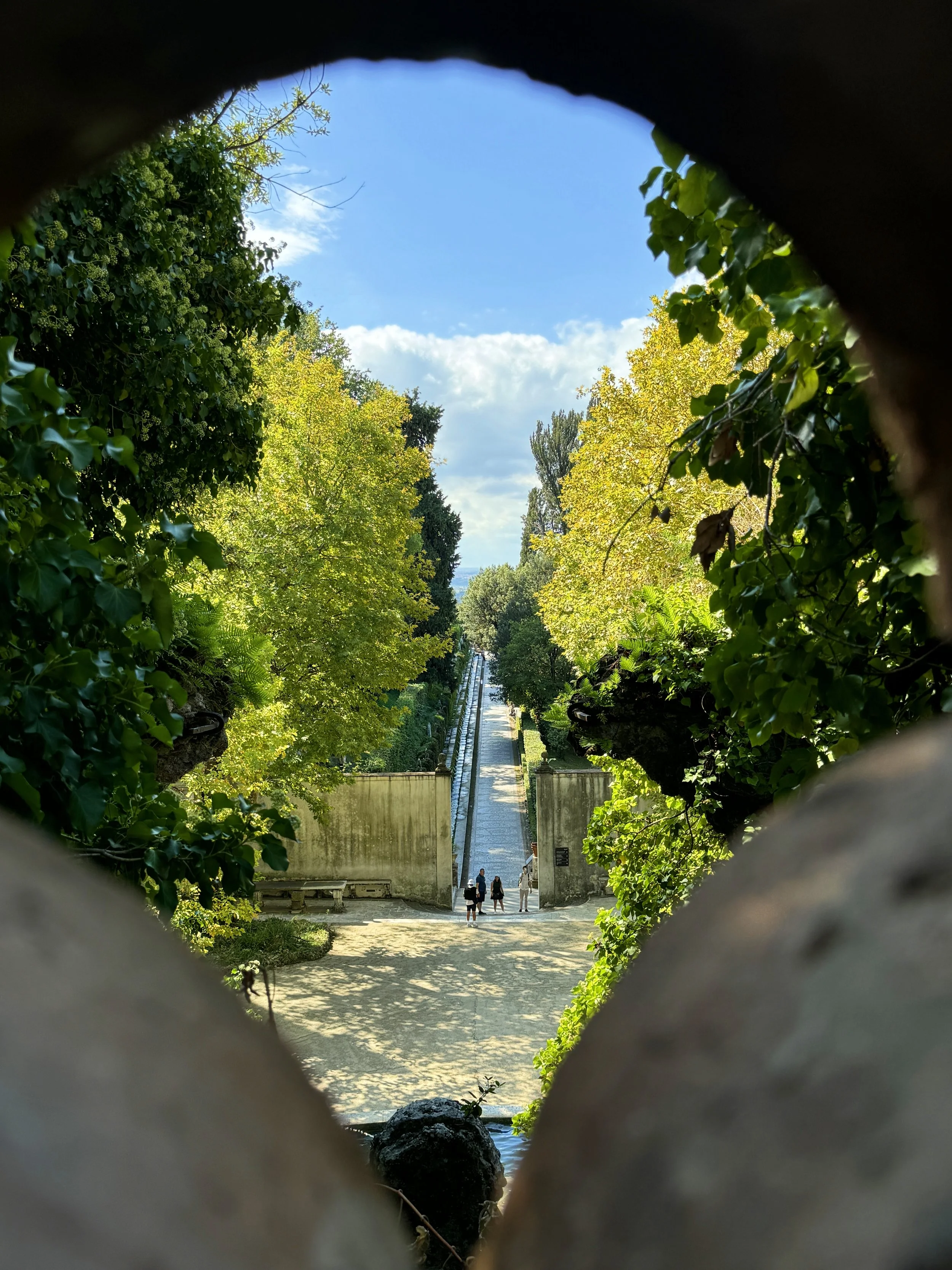 People walking near a fountain with a long water feature, viewed through a hole in a stone structure surrounded by greenery, trees, and a blue sky.