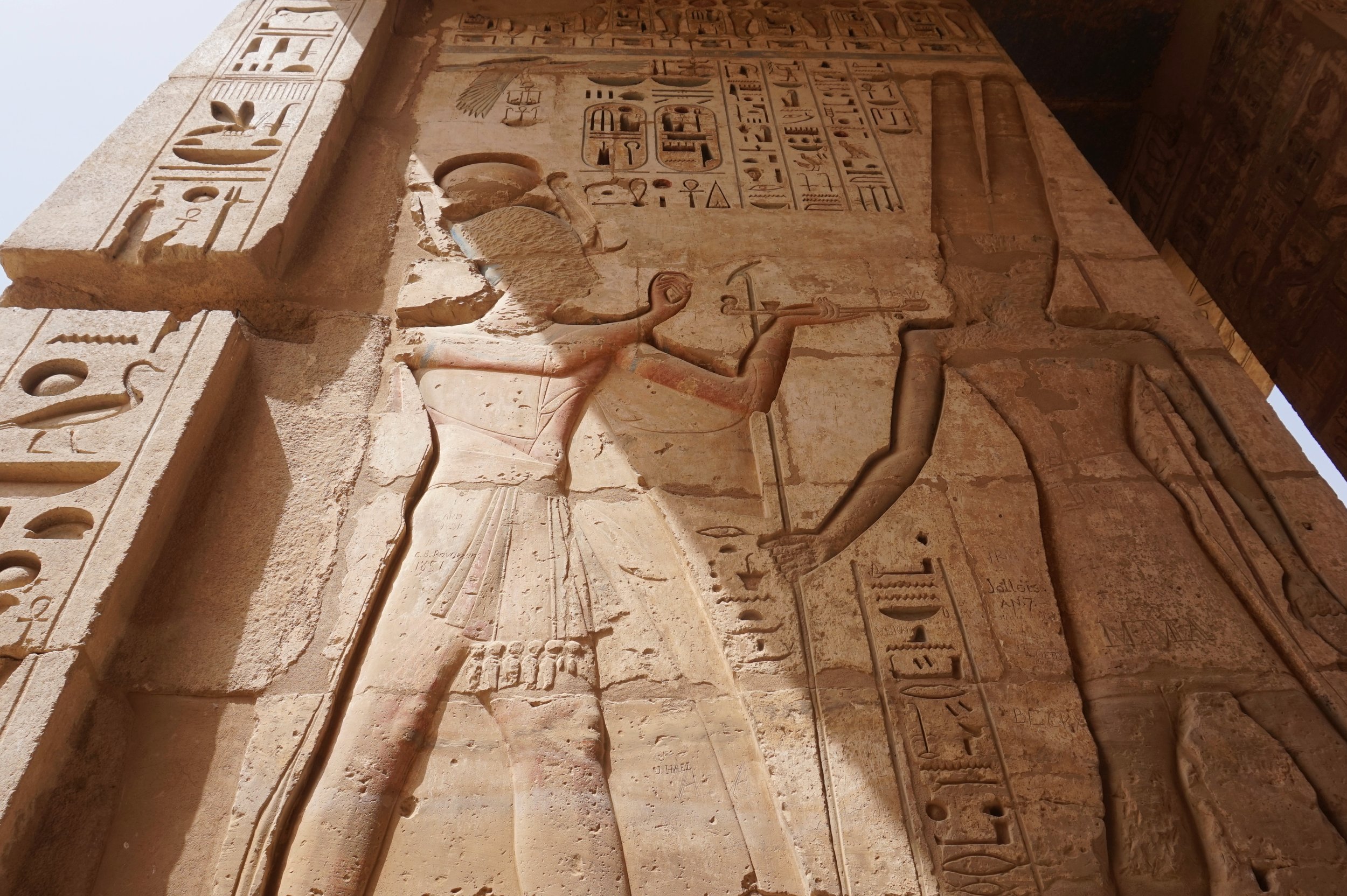 Ancient Egyptian hieroglyphs and carvings on a large stone wall, featuring various symbols, figures, and inscriptions.