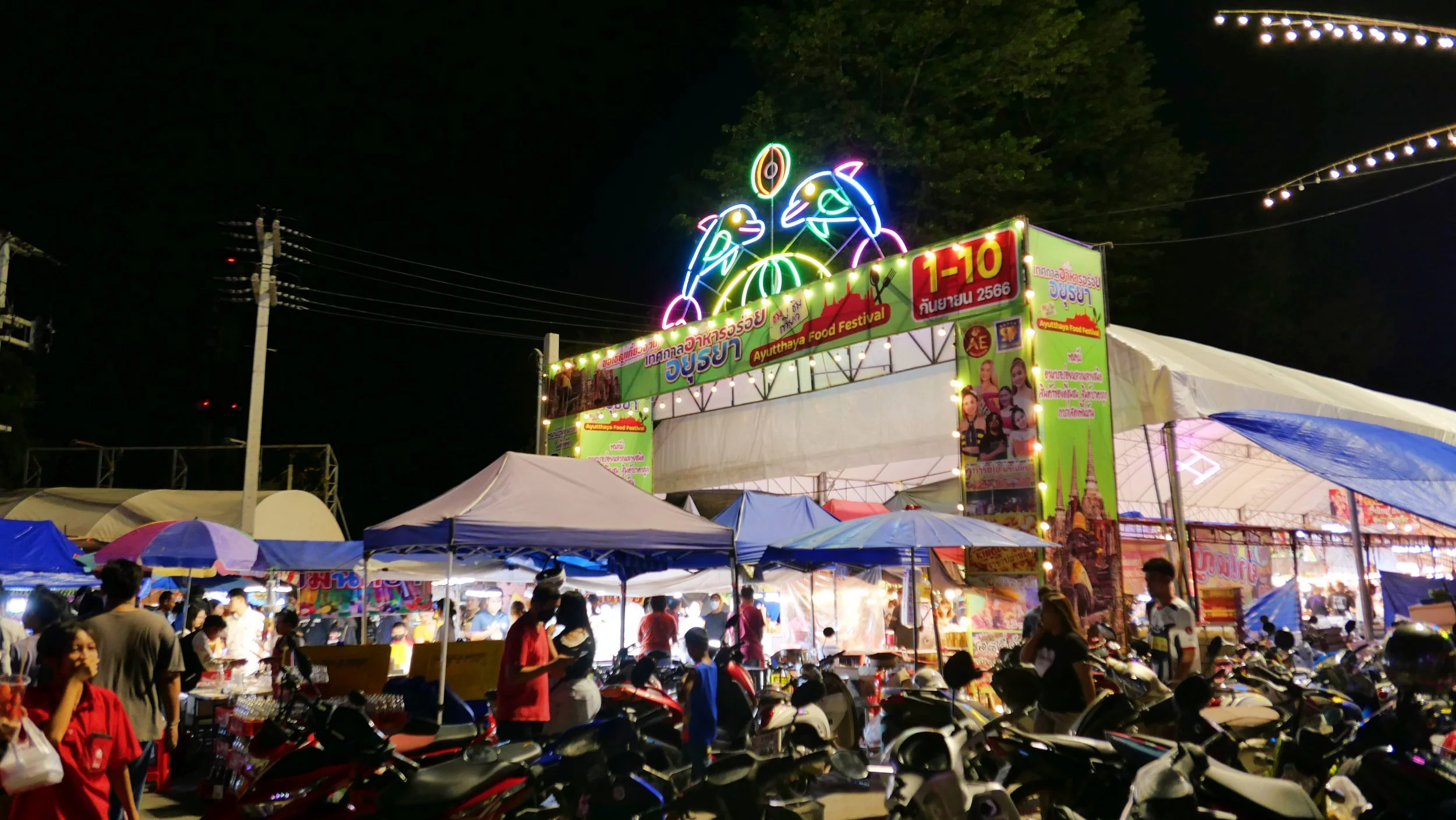Night scene at a lively festival with numerous tents, many people, and neon signs. A prominent sign reads 'Ayutthaya Food Festival' in Thai and English, with decorative neon lights and a crowd browsing food stalls.