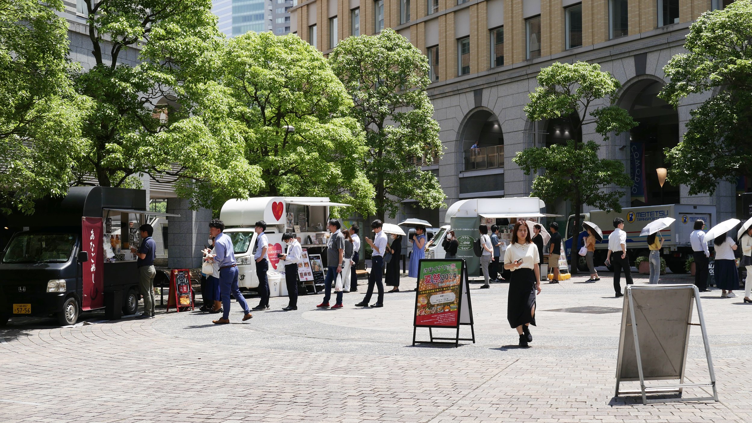 People walking and queuing at food trucks in an urban street, with tall buildings and green trees in the background.