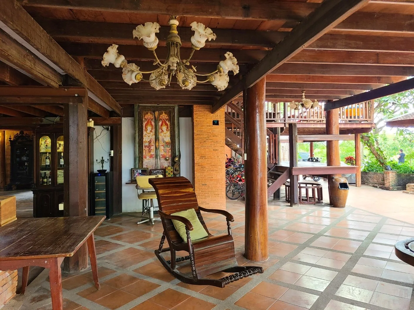 Interior view of a rustic wooden house with furniture including a wooden rocking chair with a pillow, a table, and chandeliers, with an outdoor patio and greenery visible outside.