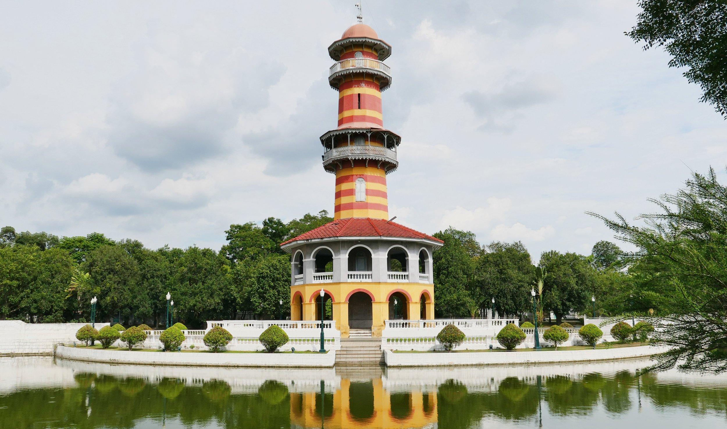 A colorful, striped tower with multiple levels, surrounded by greenery and a small body of water in the foreground.