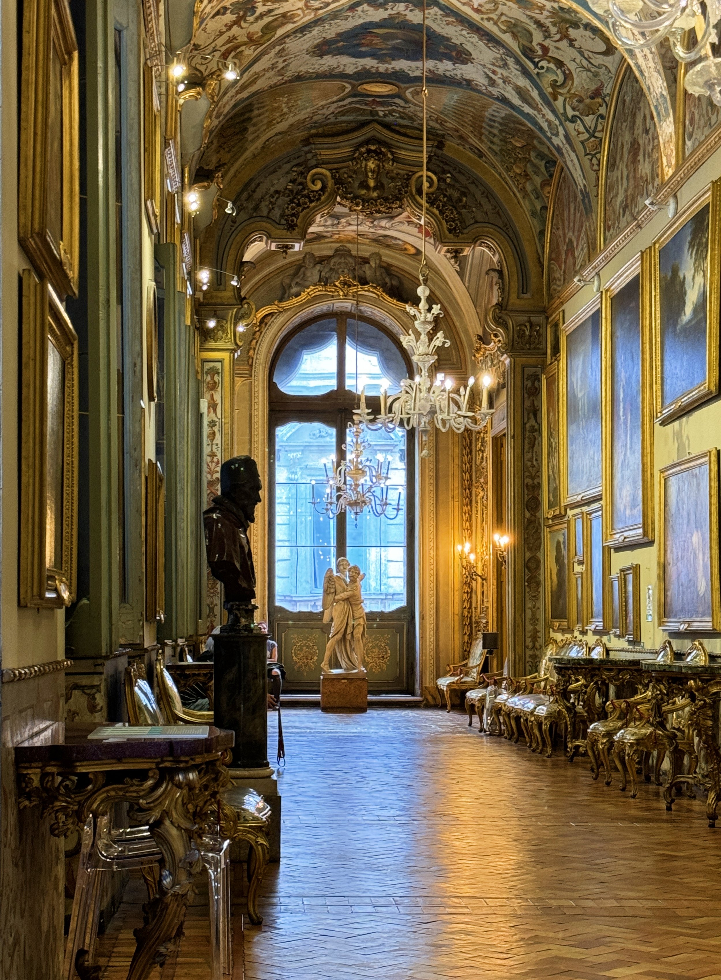 An ornate hallway with gilded moldings, chandeliers, and classical statues, illuminated by warm lighting, with a large window at the end of the hall showing an outdoor scene.